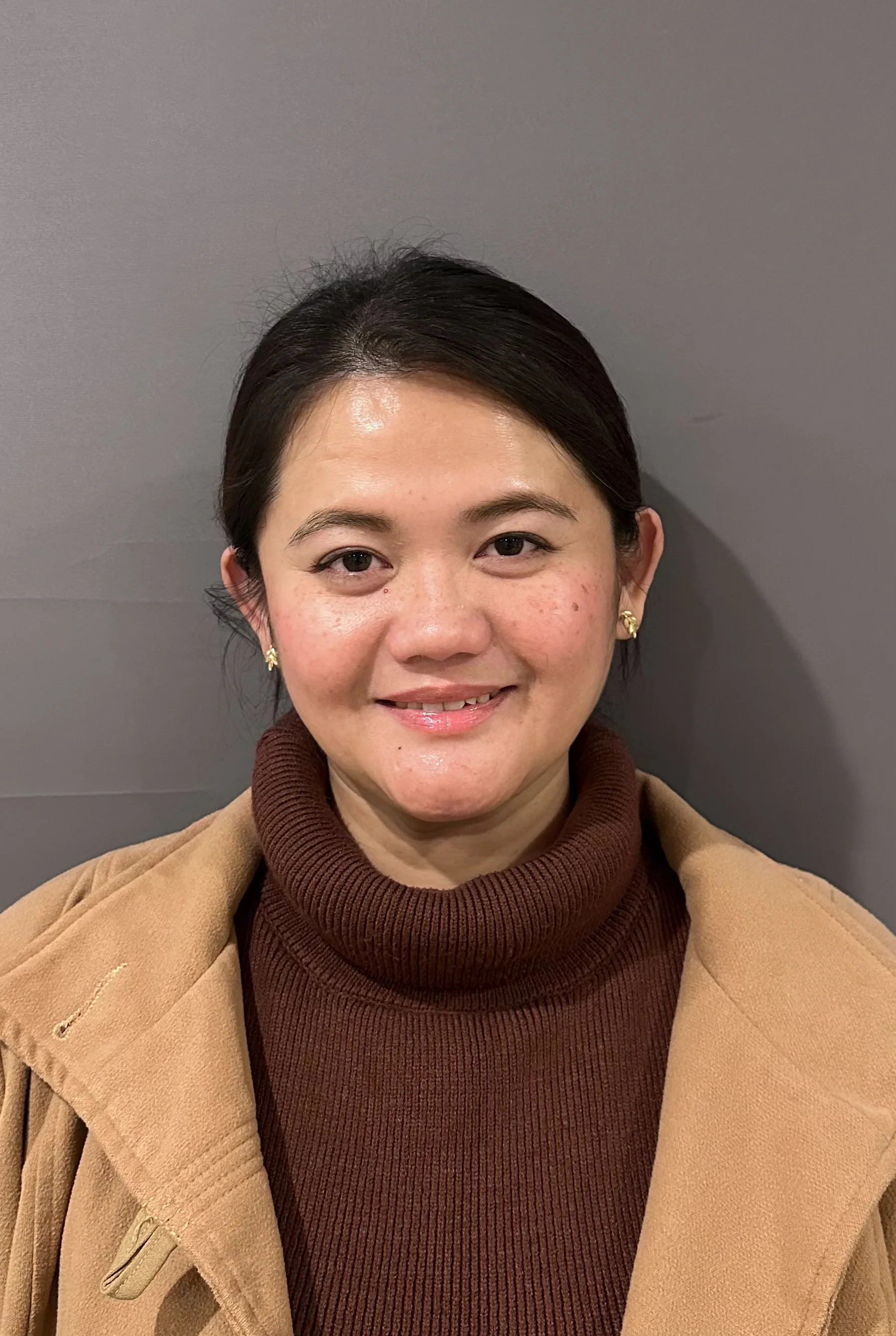 Close-up of a smiling woman with dark hair, wearing a brown knit turtleneck and a tan coat, standing against a gray wall.