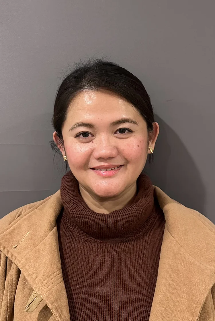 Close-up of a smiling woman with dark hair, wearing a brown knit turtleneck and a tan coat, standing against a grey wall.