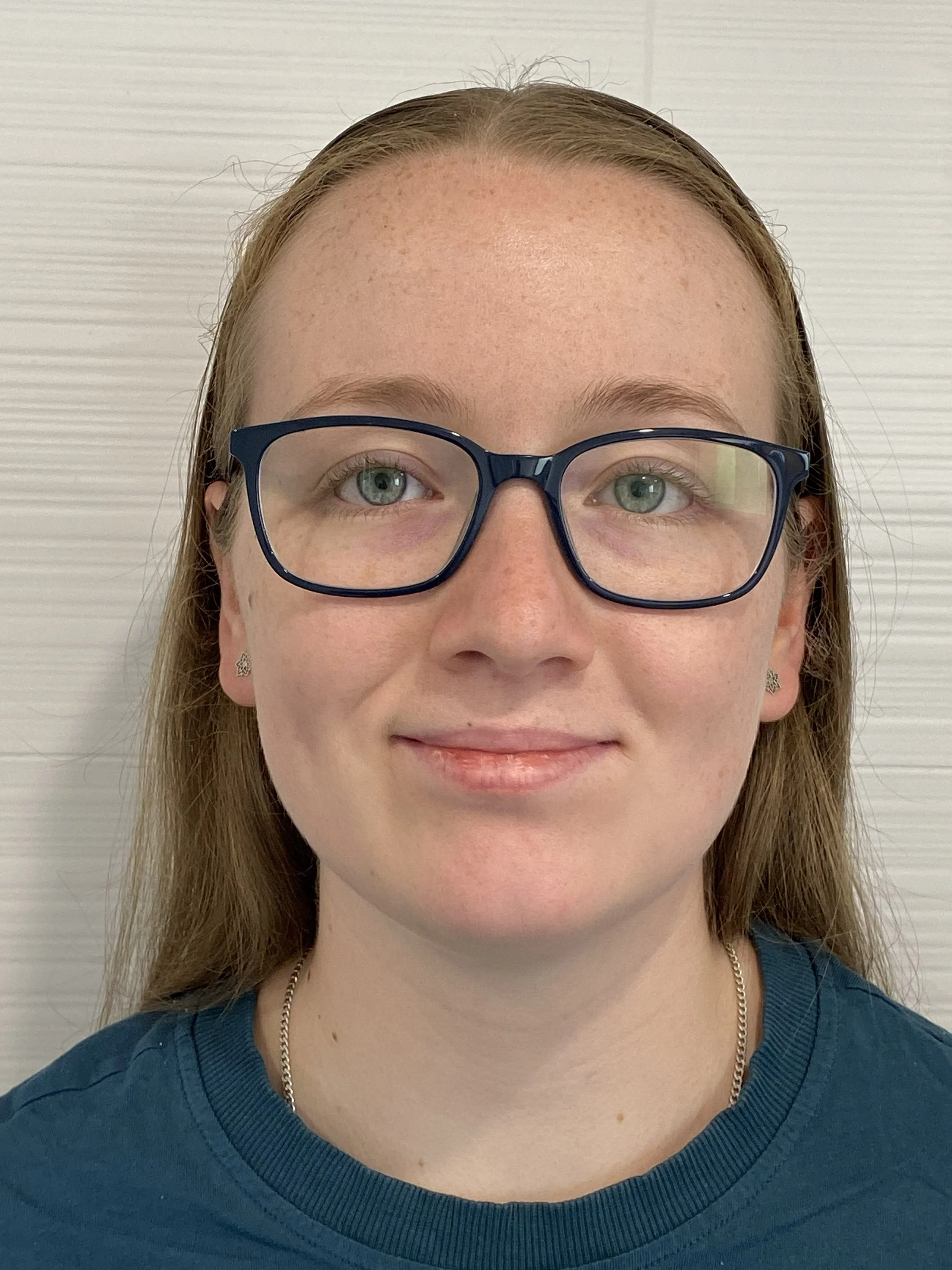 Close-up of a woman with glasses, blue shirt, and a necklace, smiling slightly against a light-colored background.