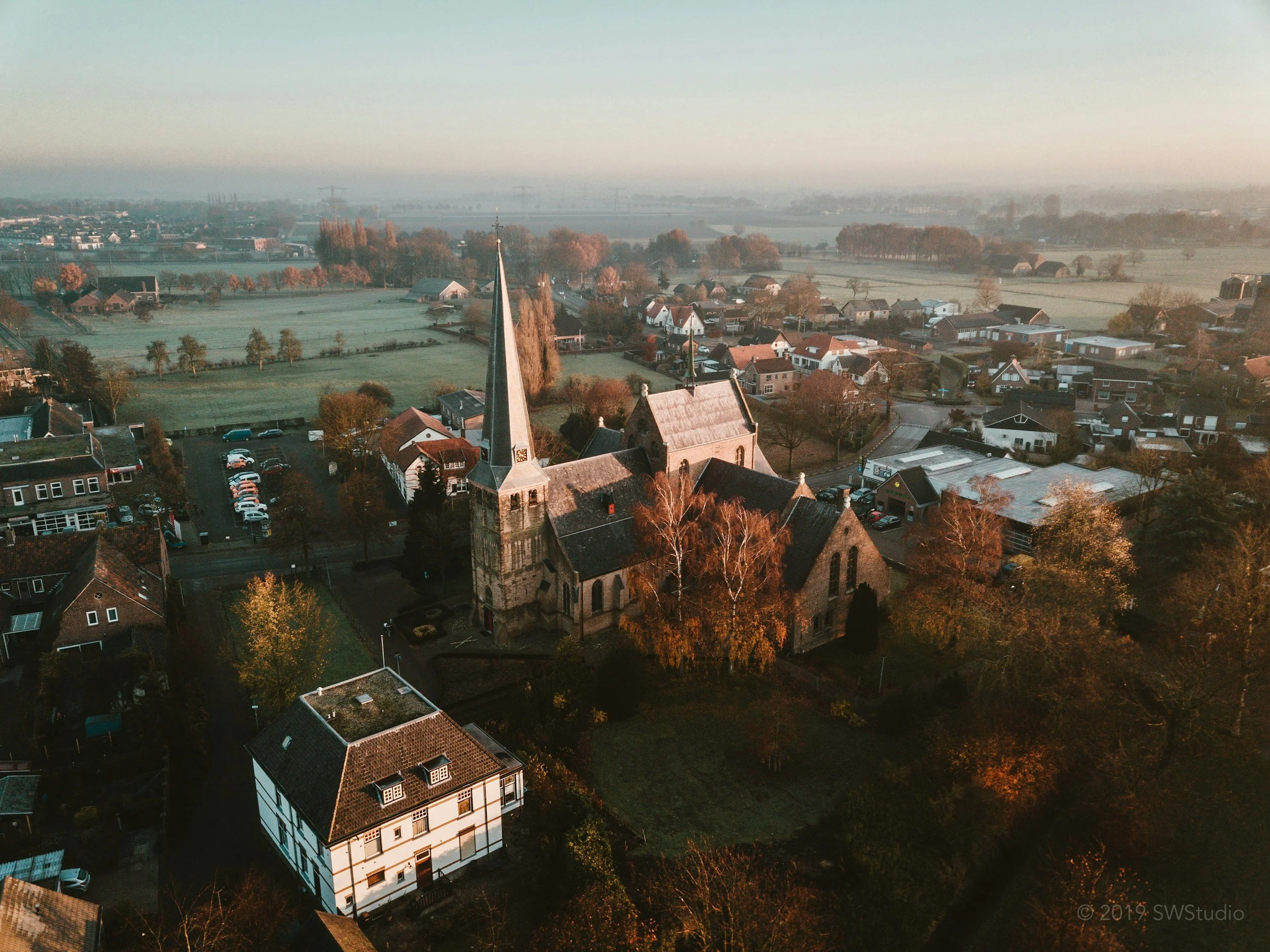 Aerial view of a town with a church steeple in the foreground, surrounded by houses, trees, and green fields during early morning or late afternoon sunlight.