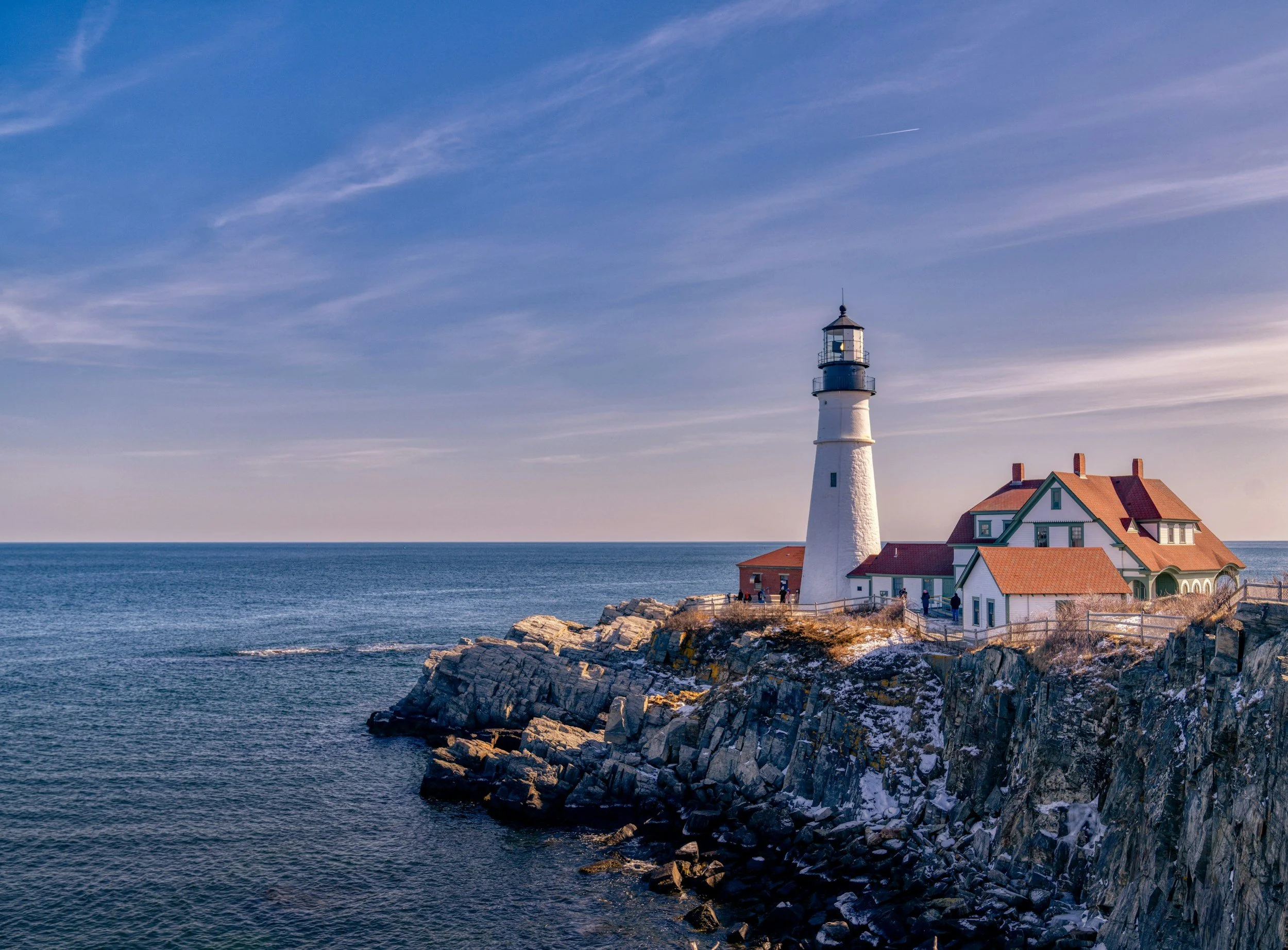 A lighthouse near Portland Maine on the ocean with rocky shoreline and a building with orange roof next to the lighthouse.