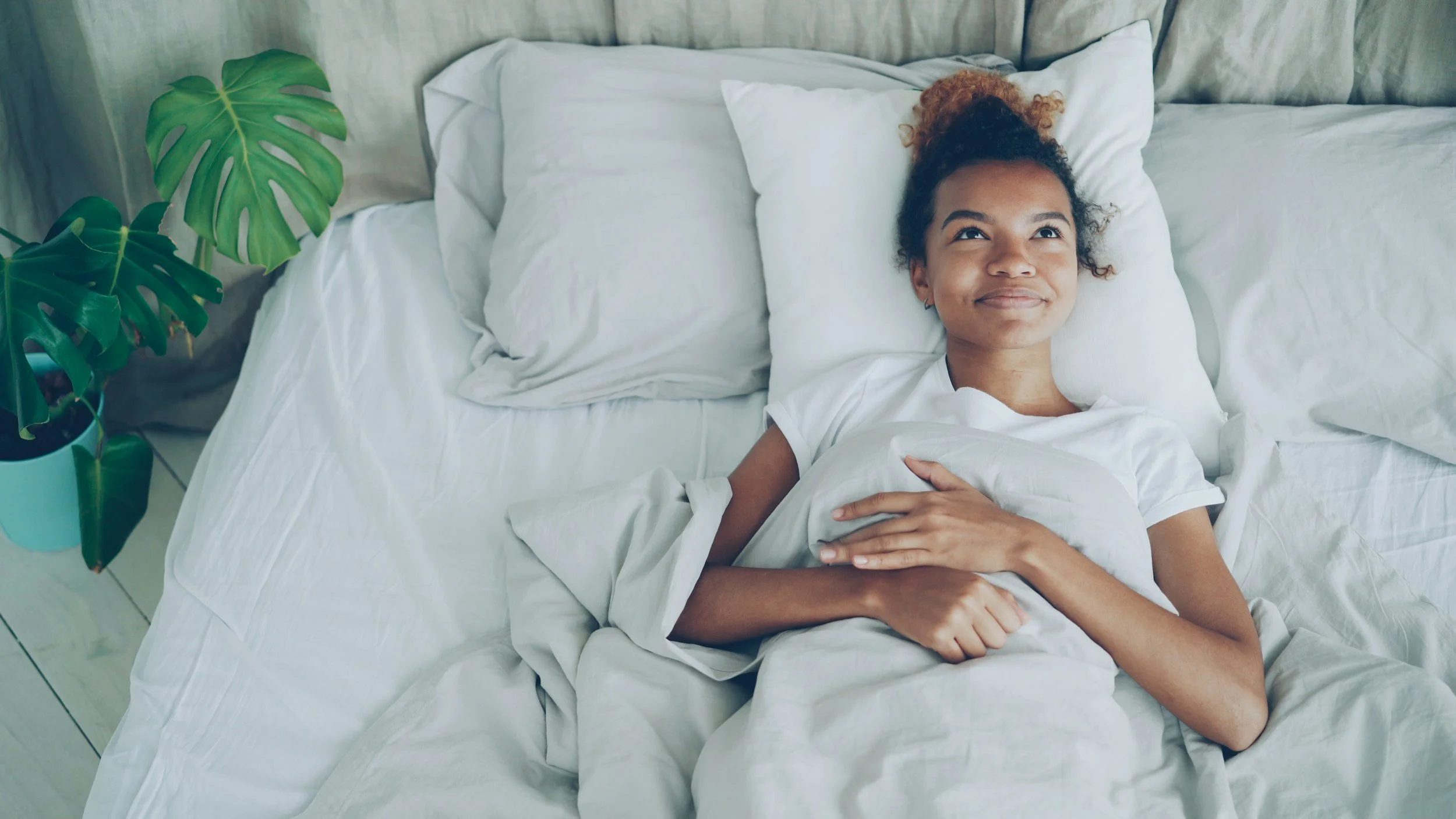 A young woman with curly hair lying in bed with white sheets, smiling and looking up, with a potted Monstera plant nearby.