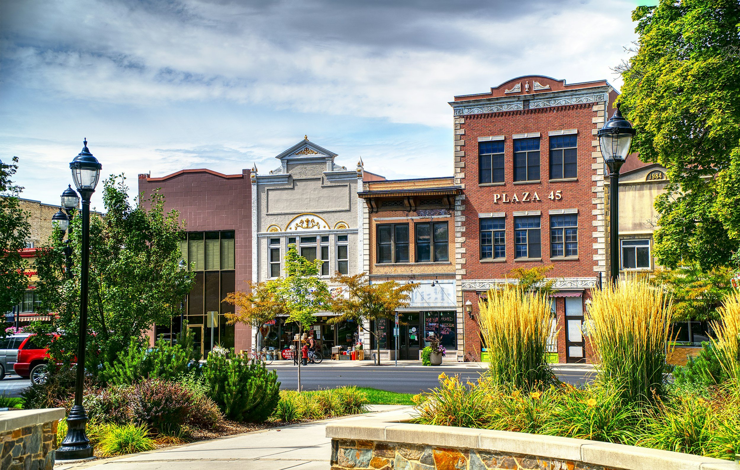 A street view with historic brick buildings and storefronts, trees, street lamps, and a park or sidewalk area with plants and flowers.