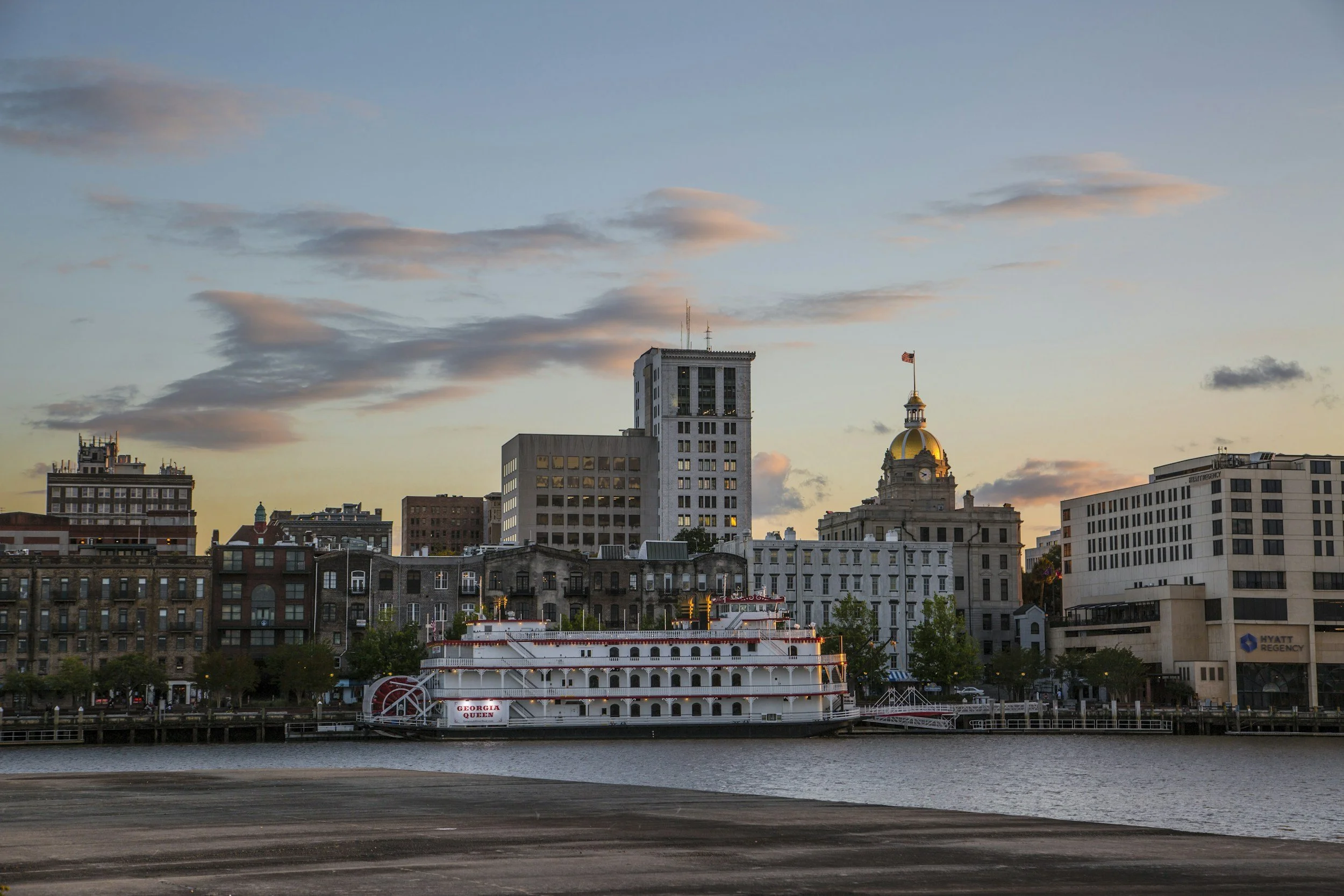Savannah, Georgia skyline at sunset with historic and modern buildings, a riverfront, and a boat named Georgia Queen in the foreground.