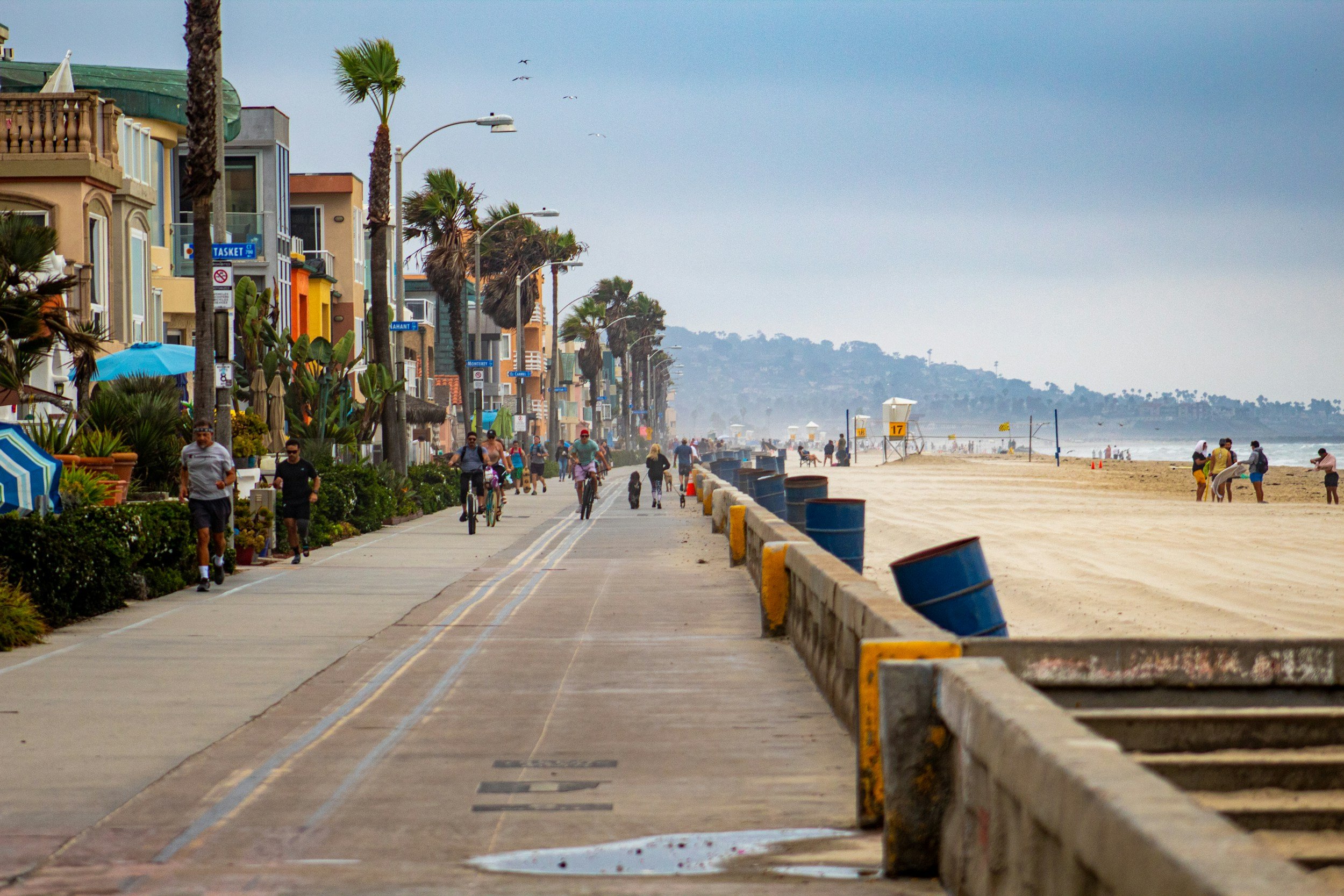 People walking and biking on a seaside promenade in San Diego, California lined with colorful buildings, palm trees, and street lamps, with a sandy beach and ocean in the background.