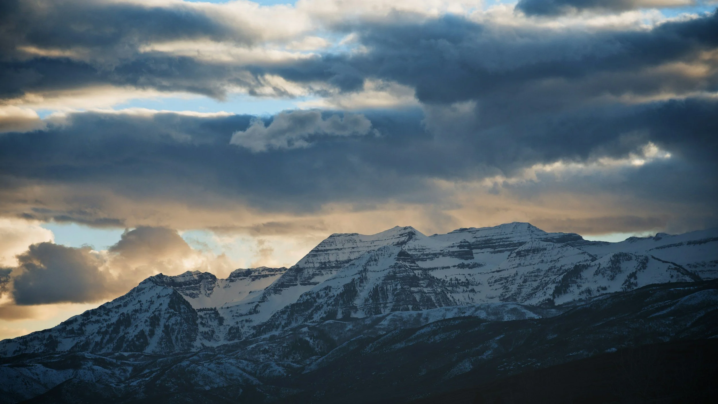 View from Midway, Utah of snow-capped mountain range under a cloudy sky with patches of sunlight breaking through.
