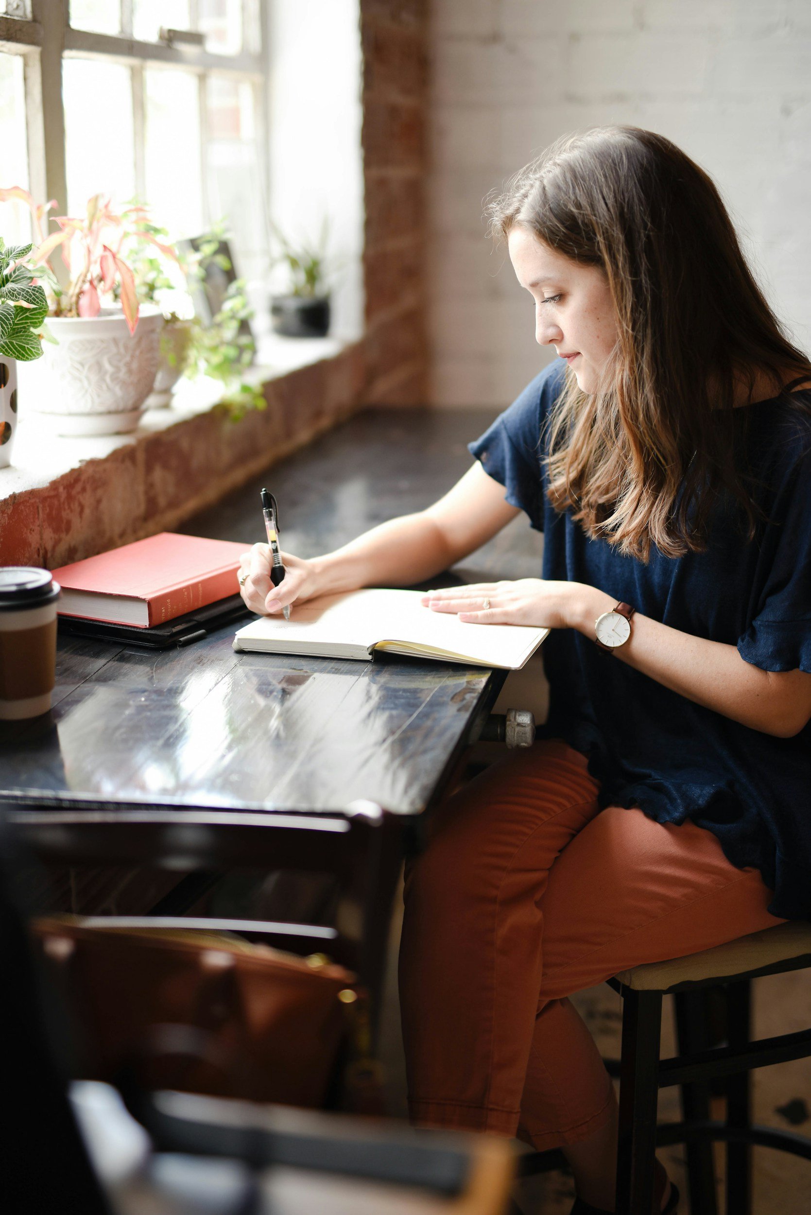 A woman with long brown hair, wearing a navy blue top, a watch, and rust-colored pants, sits at a wooden table by a window with potted plants, writing in a notebook with a pen.