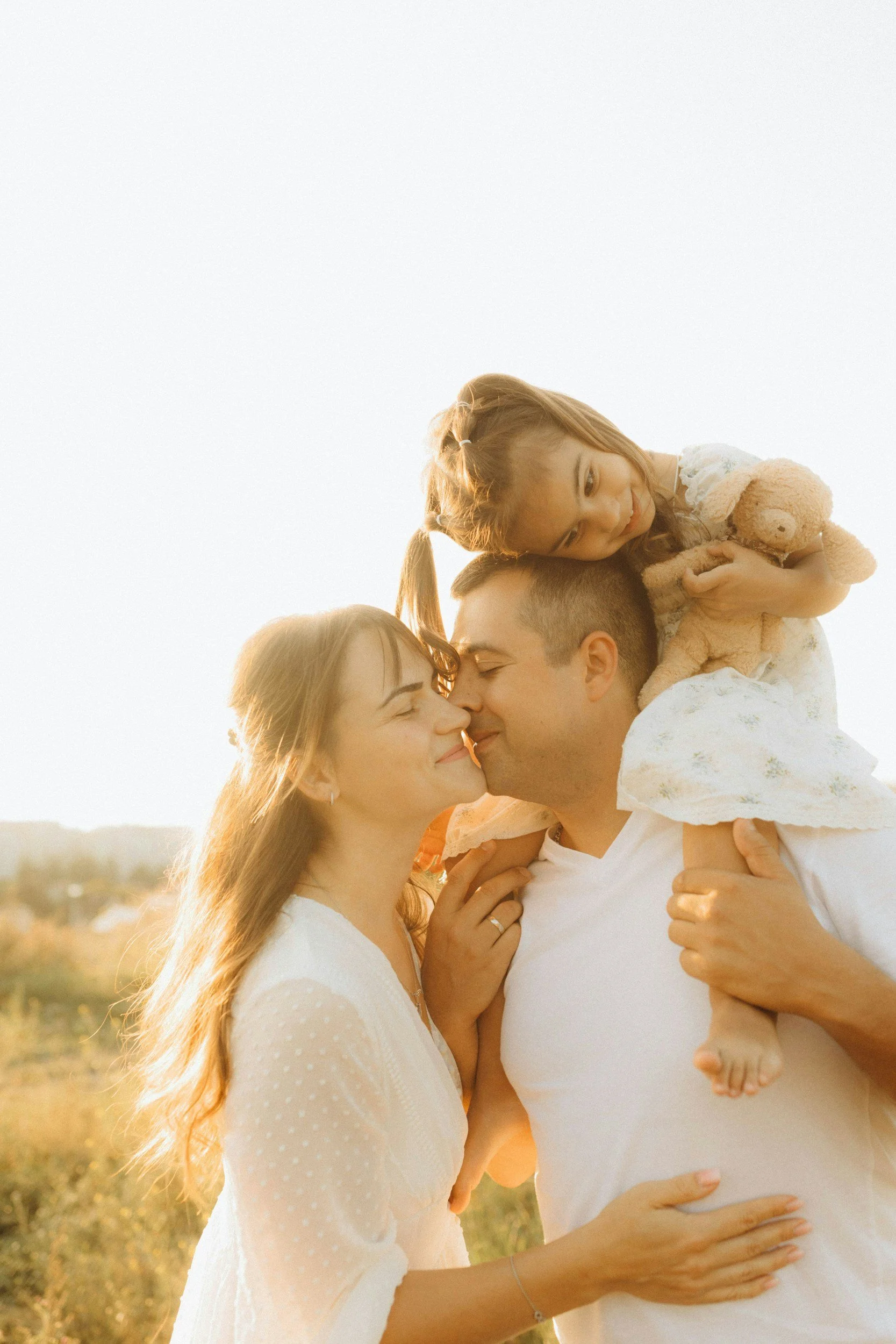 A family of three, a man, a woman, and a young girl, sharing a tender moment outdoors during sunset. They are smiling, with their faces close, and the girl is holding a teddy bear while sitting on the man's shoulders. The scene is warm and loving.