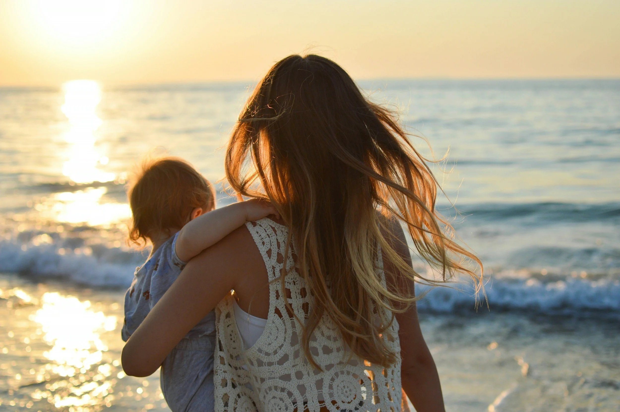 A woman with long hair is holding a small child as they watch the sunset at the beach, with ocean waves in the background.