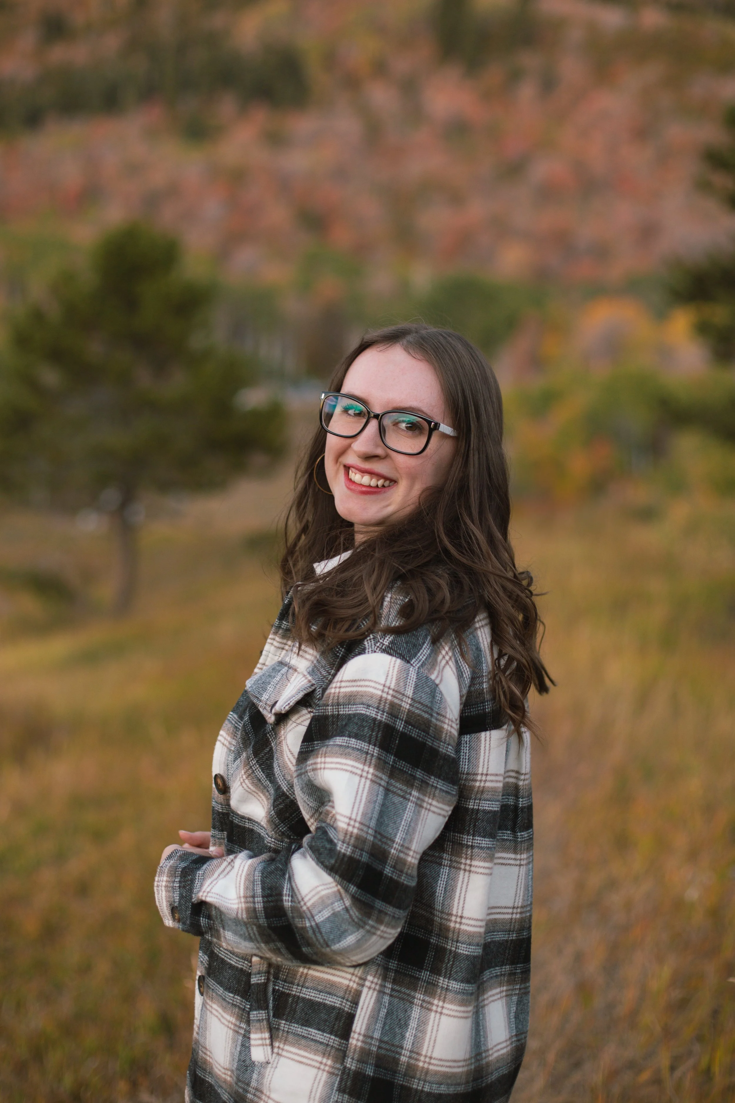 A young woman with long brown hair, wearing glasses and a plaid coat, smiling outdoors with autumn trees in the background.