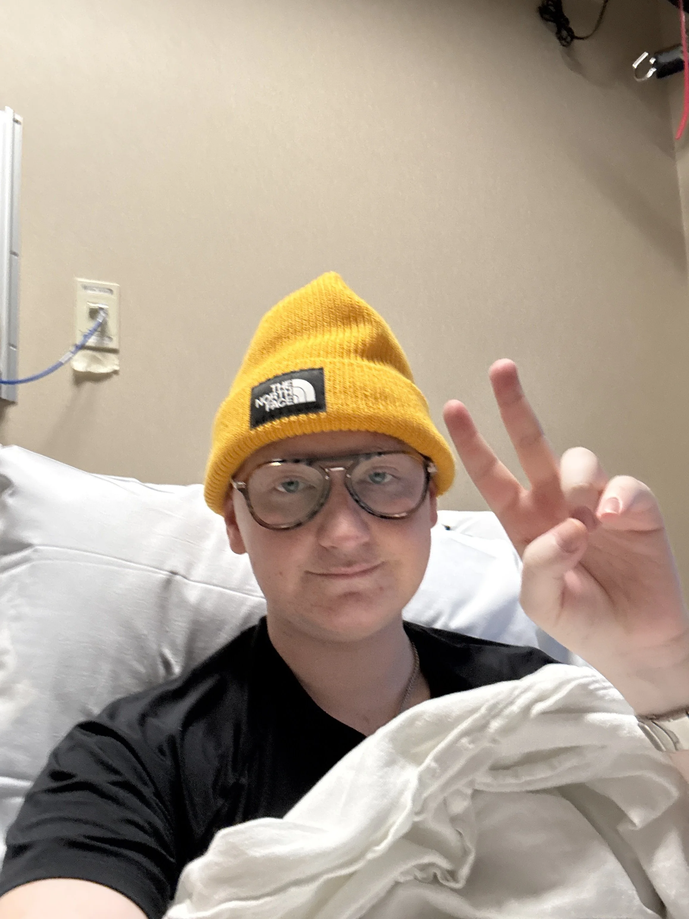 Bear wearing glasses and a bright yellow beanie taking a selfie in a hospital room, making a peace sign with their right hand.