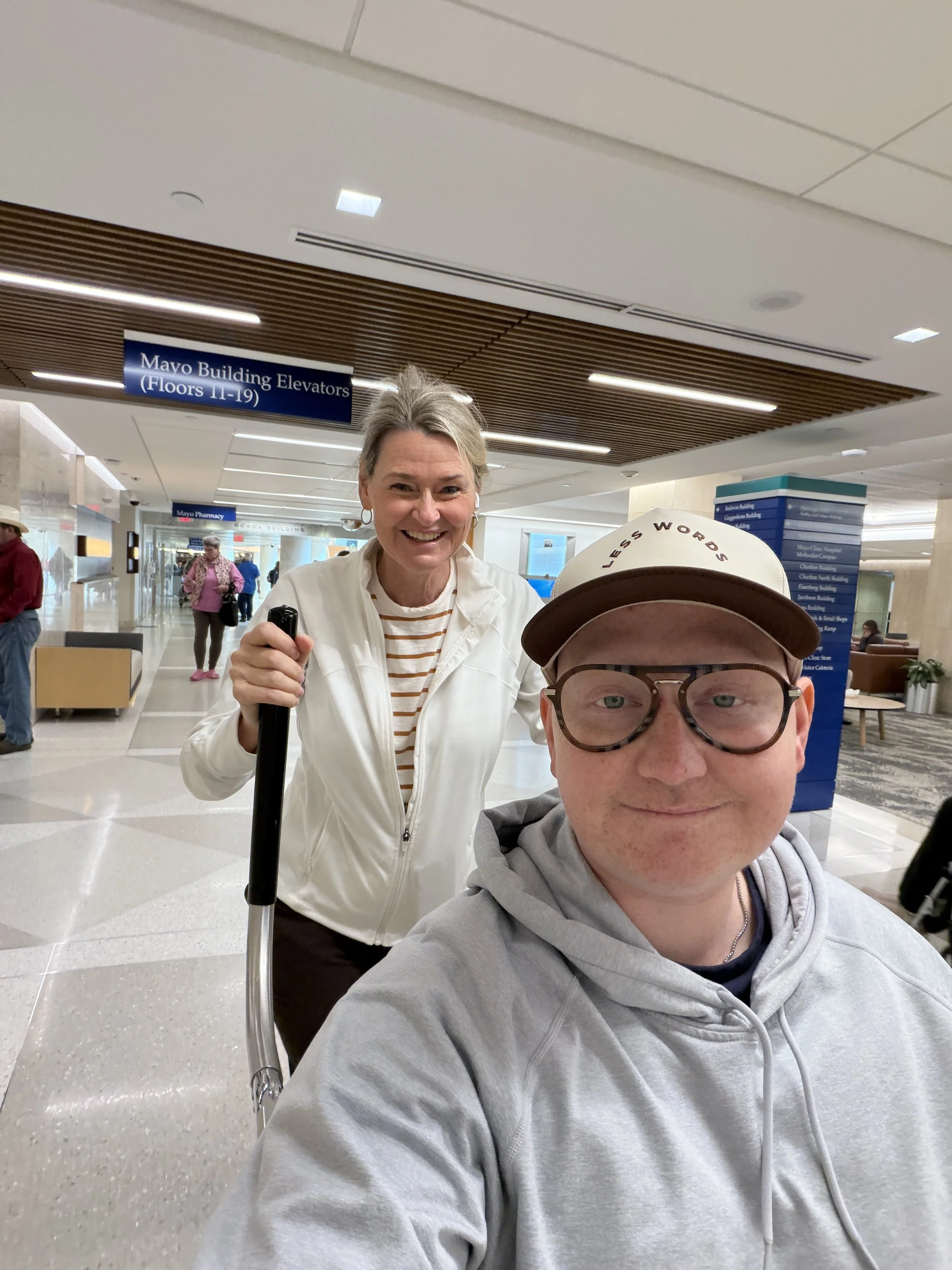 Bear and Ann. Ann is holding a device and wearing a white jacket, while the man is wearing a gray hoodie and a beige cap with the words 'Less Words'. They are at Mayo Clinic.