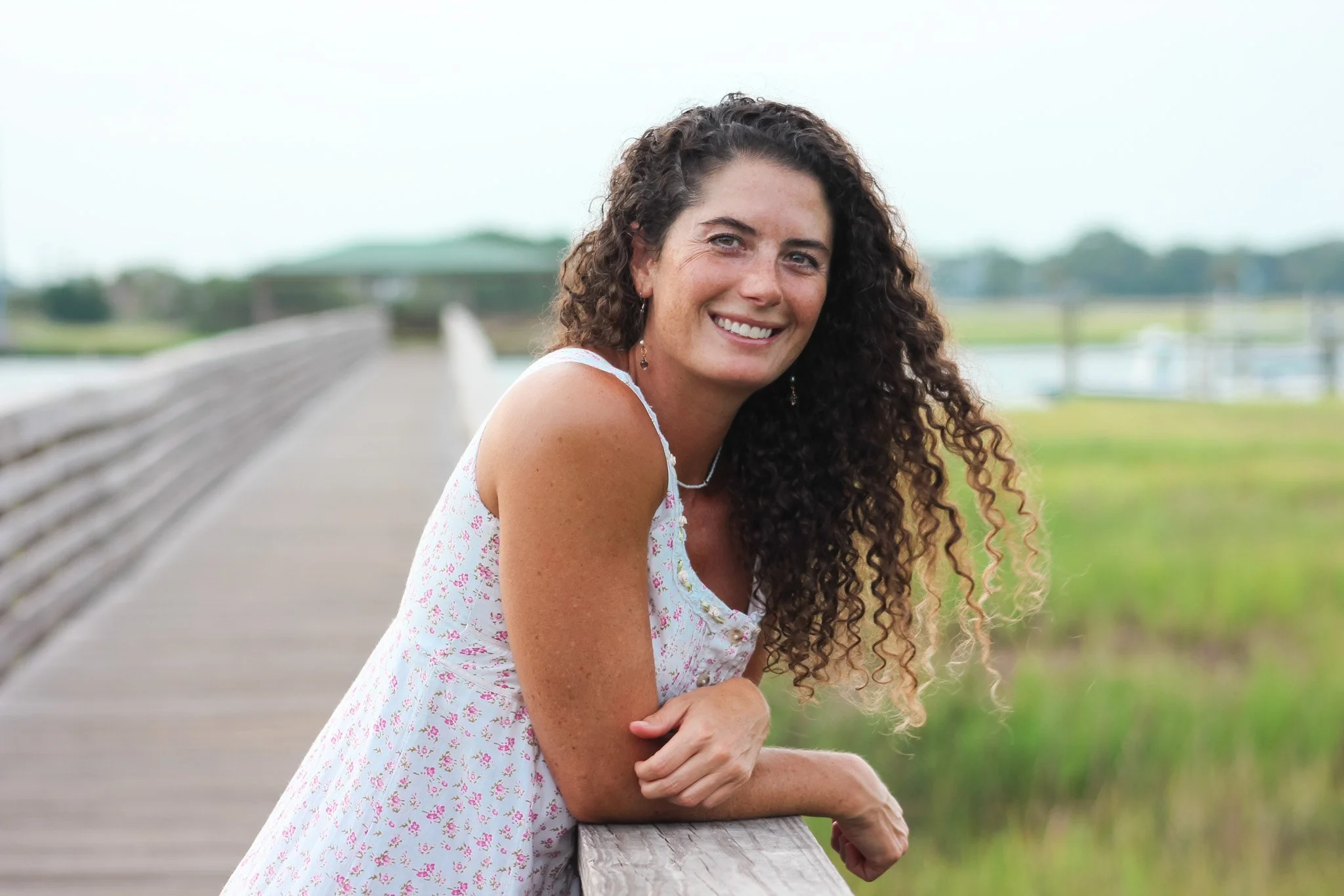 A woman with long curly hair sitting outdoors, smiling, leaning on a wooden railing at a park or trail.