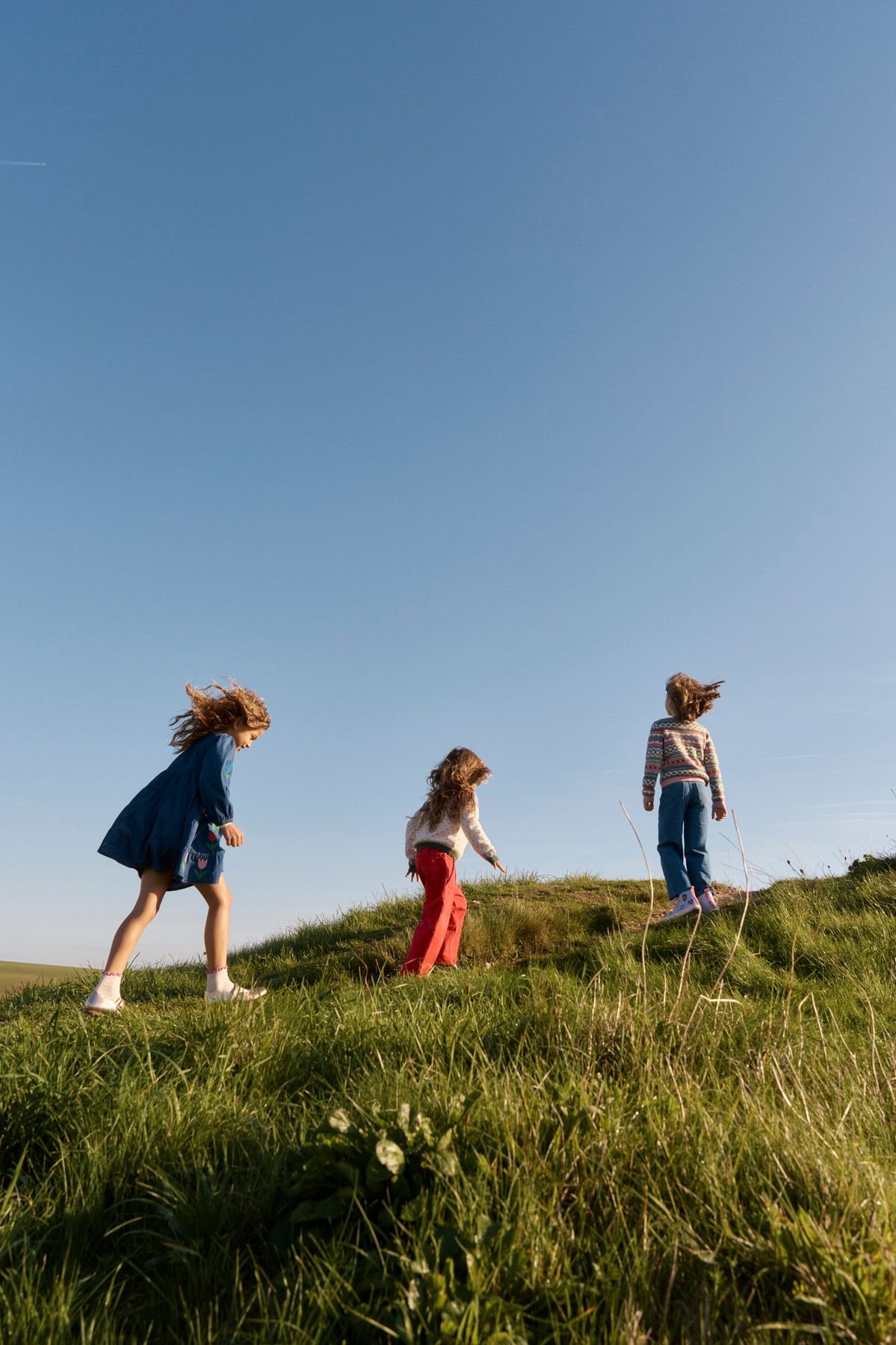 Three children playing on a grassy hill under a clear blue sky.