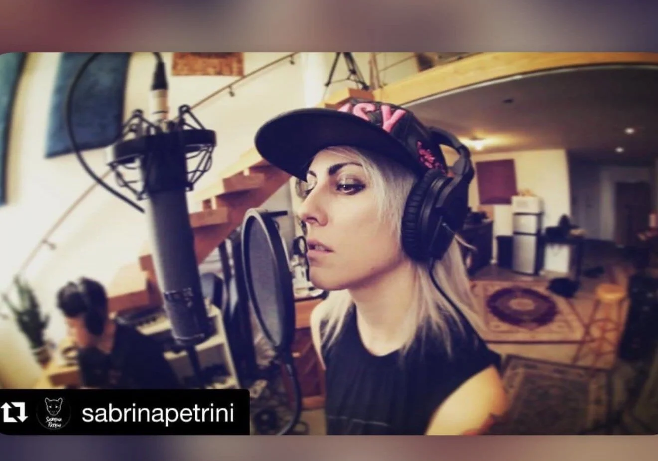 Woman with blonde hair wearing a black hat and headphones singing or speaking into a microphone in a recording studio with a man in the background working at a computer.