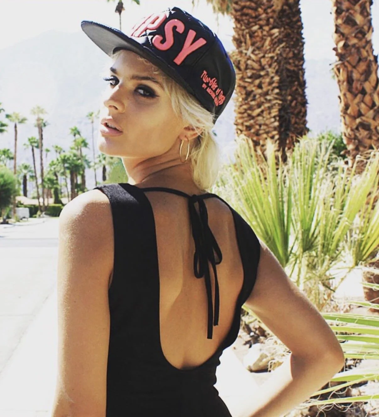 A woman with blonde hair wearing a black sleeveless dress with an open back and a black baseball cap with pink text, standing outdoors near palm trees.