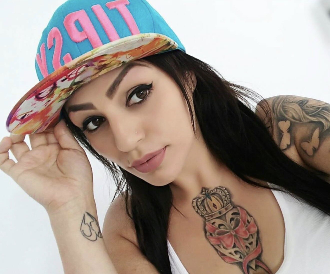 Woman with long black hair and tattoos, wearing a colorful hat that says 'LIFE' in pink, posing with her hand on the brim of the hat.
