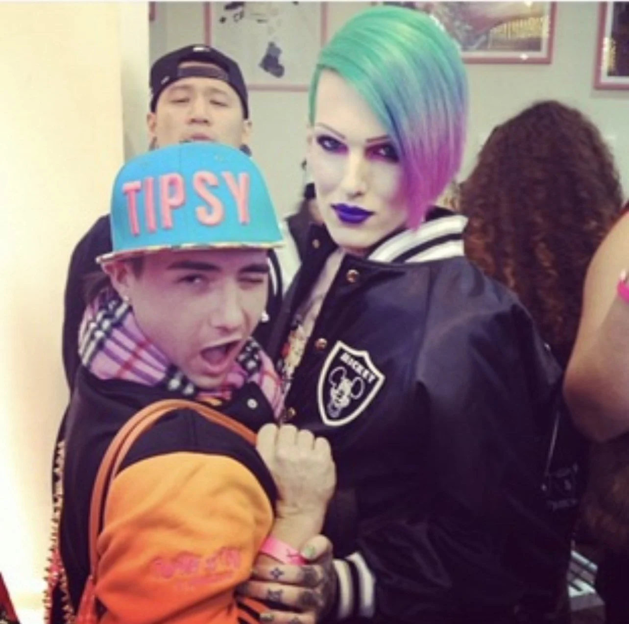 A person with rainbow-colored hair and dark makeup posing with two men at a social event. One man is wearing a colorful cap and a jacket with an orange sleeve, while the other is in the background wearing a black cap. The scene appears lively.