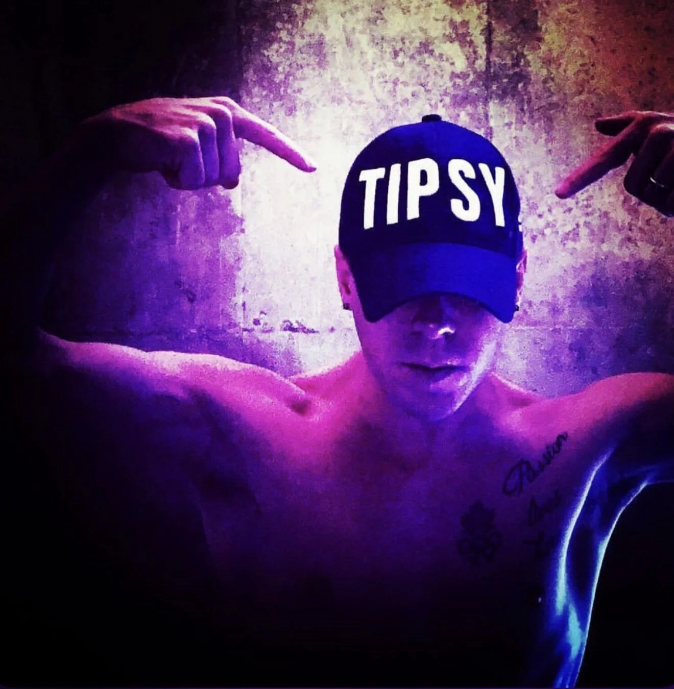 A shirtless man wearing a cap that says 'TIPSY' and pointing at his head with both hands, with tattoos on his left arm and chest, against a textured wall background with purple lighting.