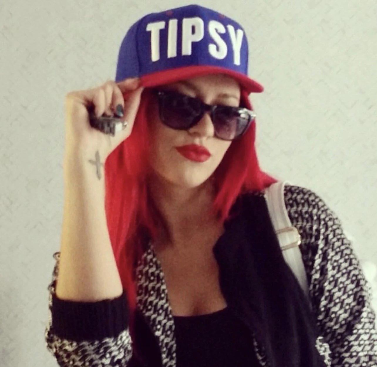 A woman with red hair wearing a blue baseball cap with 'TIPSY' written on it, black sunglasses, red lipstick, and a patterned black and white jacket.