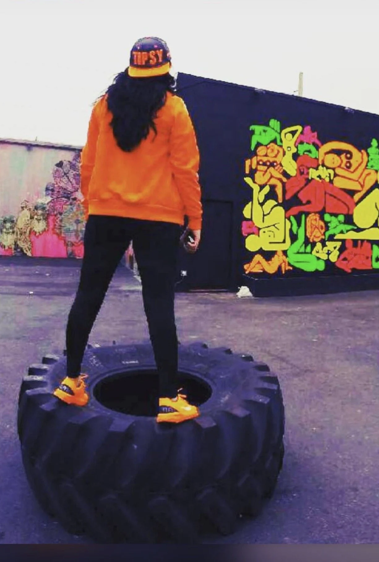 Person standing on a large black tire outdoors, facing away, wearing an orange jacket, black pants, orange shoes, and a cap that says "TIPSY" with colorful graffiti art in the background.