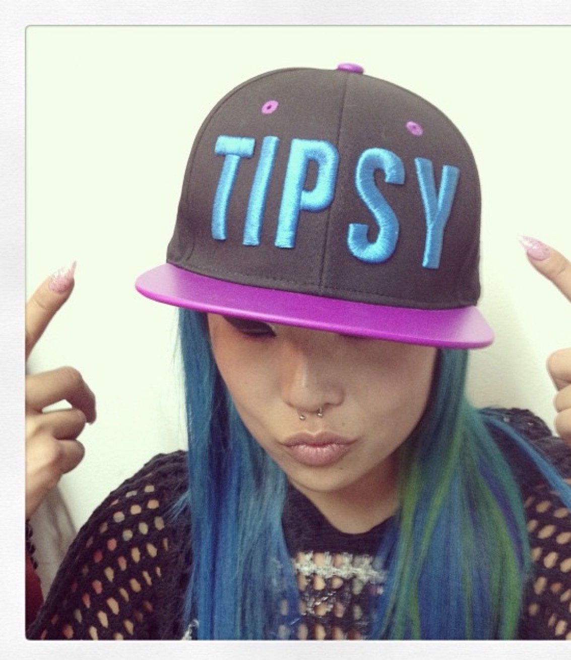 Person with blue and green hair wearing a black mesh top and a black and pink cap that says 'TIPSY' in blue letters, pointing to the cap.