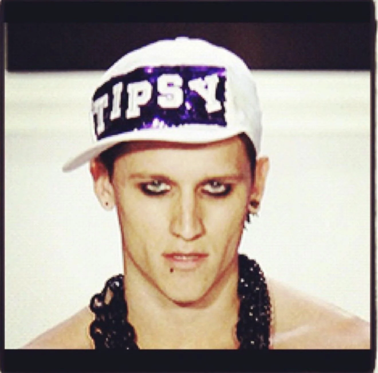 A person with makeup and a black beaded necklace wearing a white baseball cap with the word "TIPSY" written on it, tilted to the side.