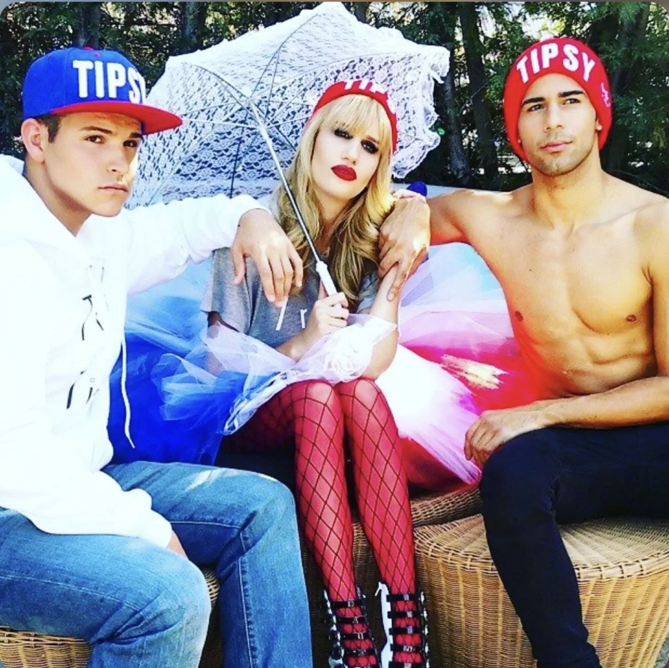 Three young adults seated outdoors on a wicker couch, with two wearing red hats that say 'TIPSY' and one holding a white lace umbrella. The woman in the middle has blonde hair, red lipstick, fishnet stockings, and a tulle skirt. The man on the right 