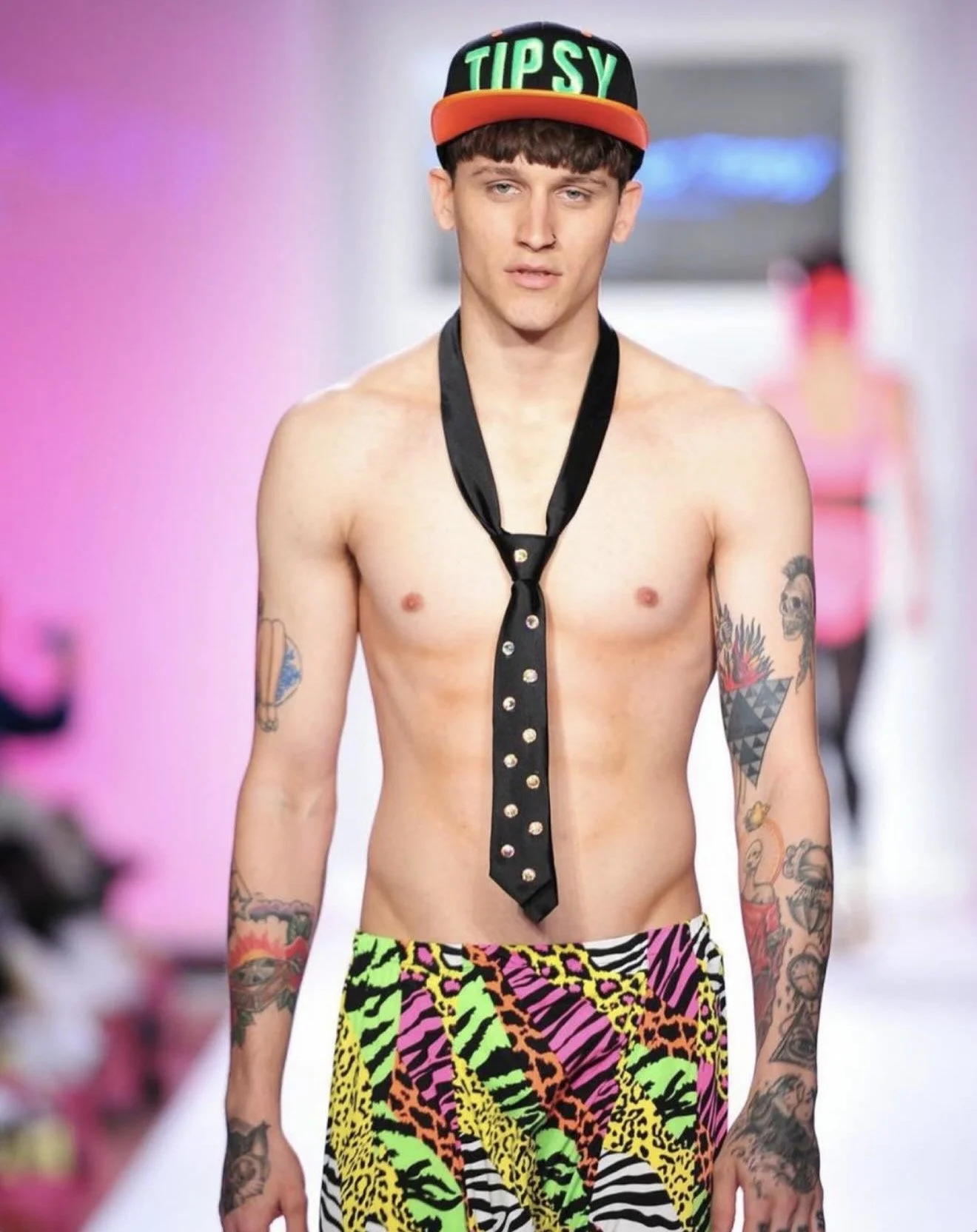 A male model with tattoos on his arms walking down a runway, wearing a black cap with green, orange, and yellow text that reads 'TIPSY,' a black tie with gold buttons, and colorful animal print shorts.