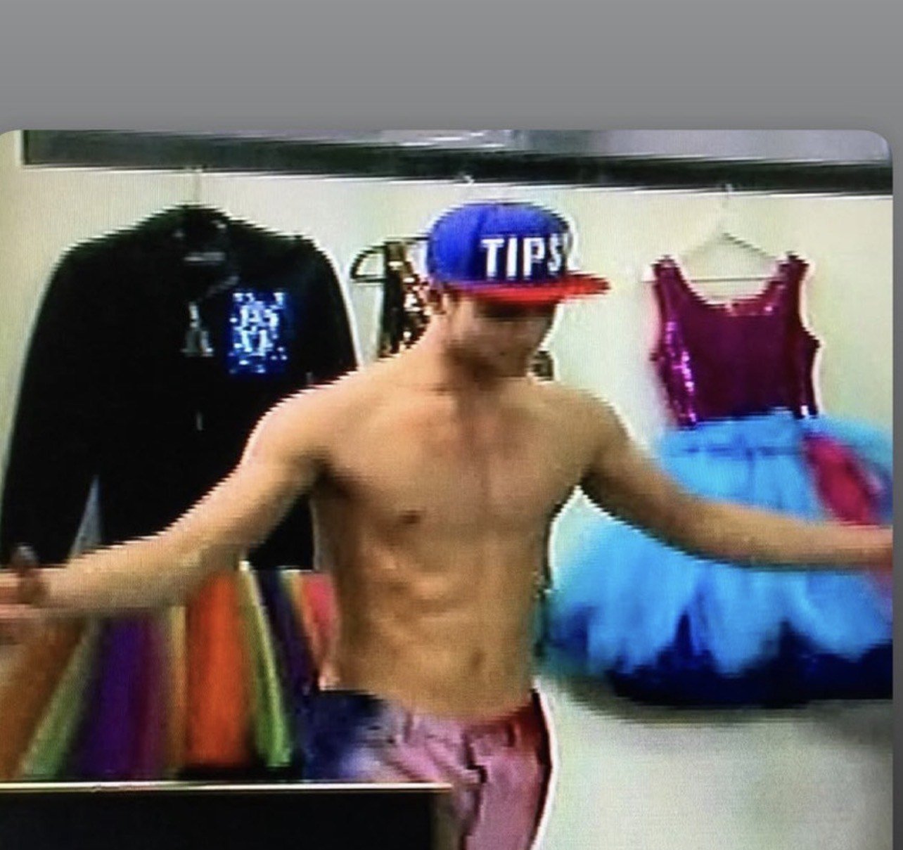 A shirtless young man wearing a blue cap with the word 'TIPS' in white and red shorts, standing in a clothing store fitting room with colorful skirts and dresses hanging behind him.