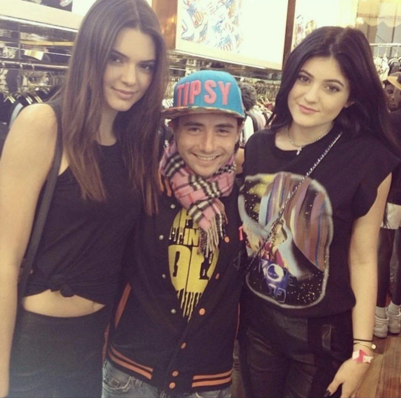Three young adults, two women and one man, smiling at the camera in a store. The man in the middle wears a colorful cap with 'TIPSY' written on it and a scarf, and has a black jacket with a yellow graphic t-shirt underneath. The women on either side 