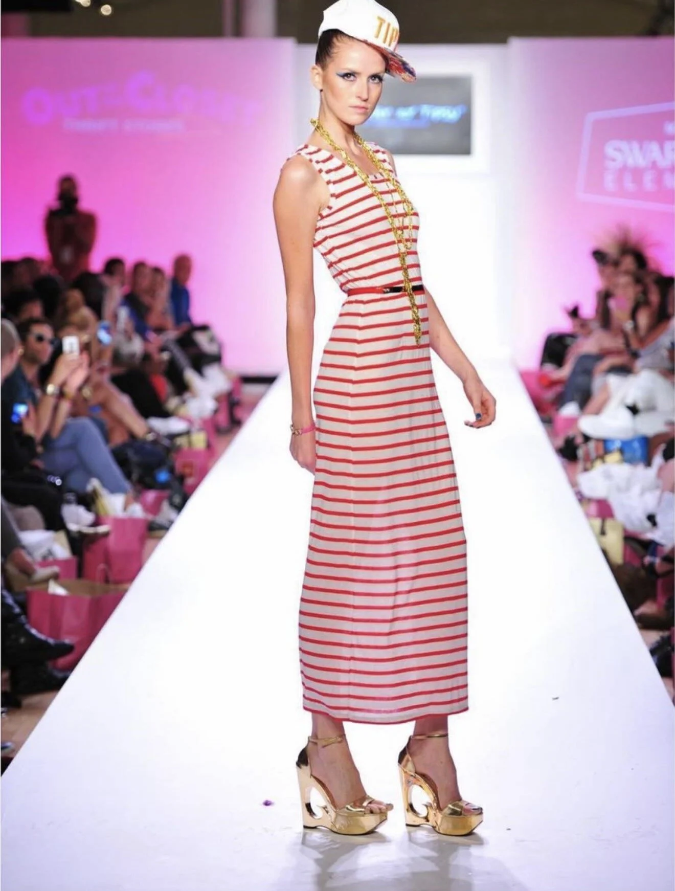 A model on a runway wearing a red and white striped maxi dress, gold platform shoes, large gold jewelry, and a white cap with red accents, during a fashion show.