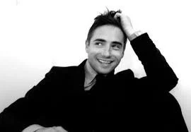 Black and white photo of a smiling man with short dark hair, wearing a dark jacket, resting his head on his hand.