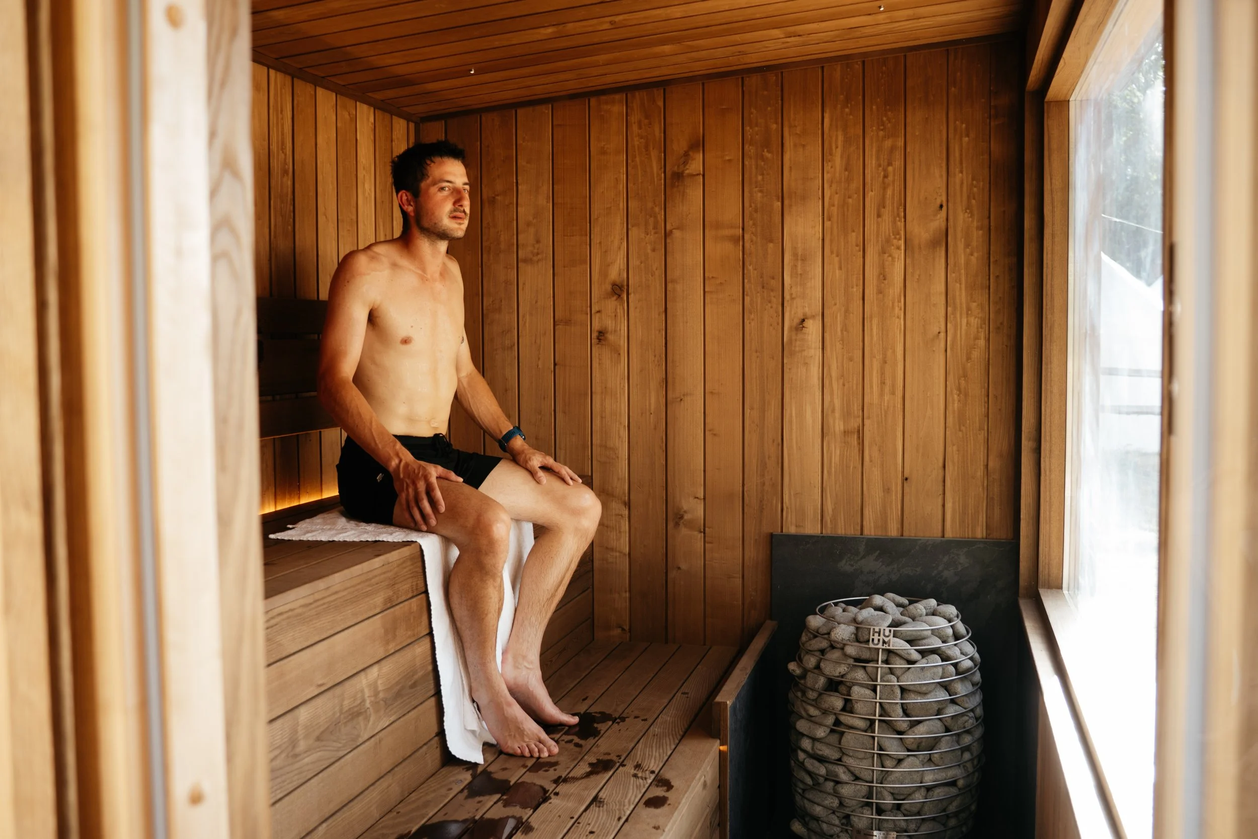Person relaxing inside The Hide sauna at Houchins, Coggeshall, with Huum heater and panoramic window overlooking the countryside.
