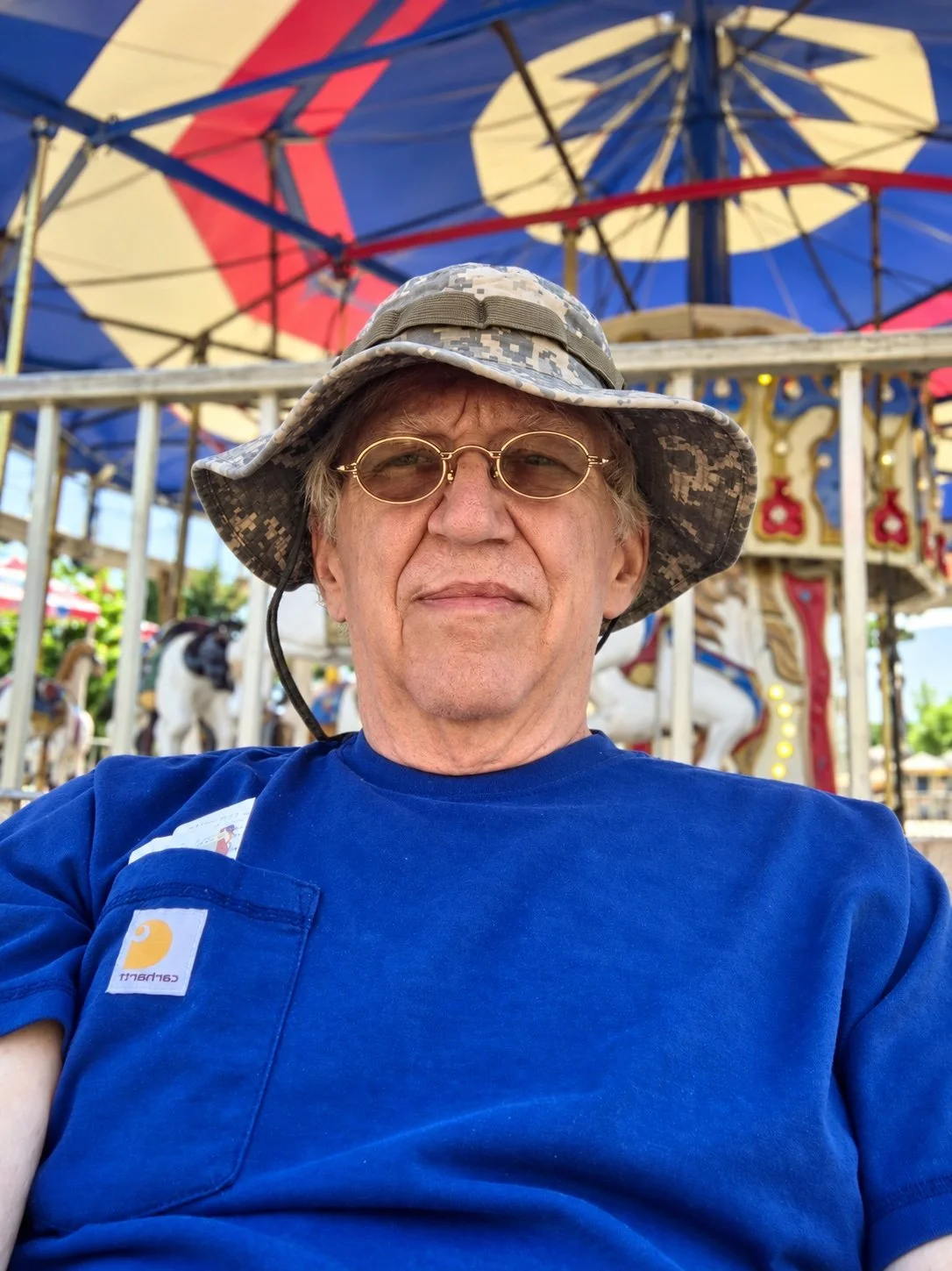An older man wearing glasses, a camouflage bucket hat, and a blue Carhartt shirt taking a selfie at a carousel amusement park ride with colorful umbrellas overhead.
