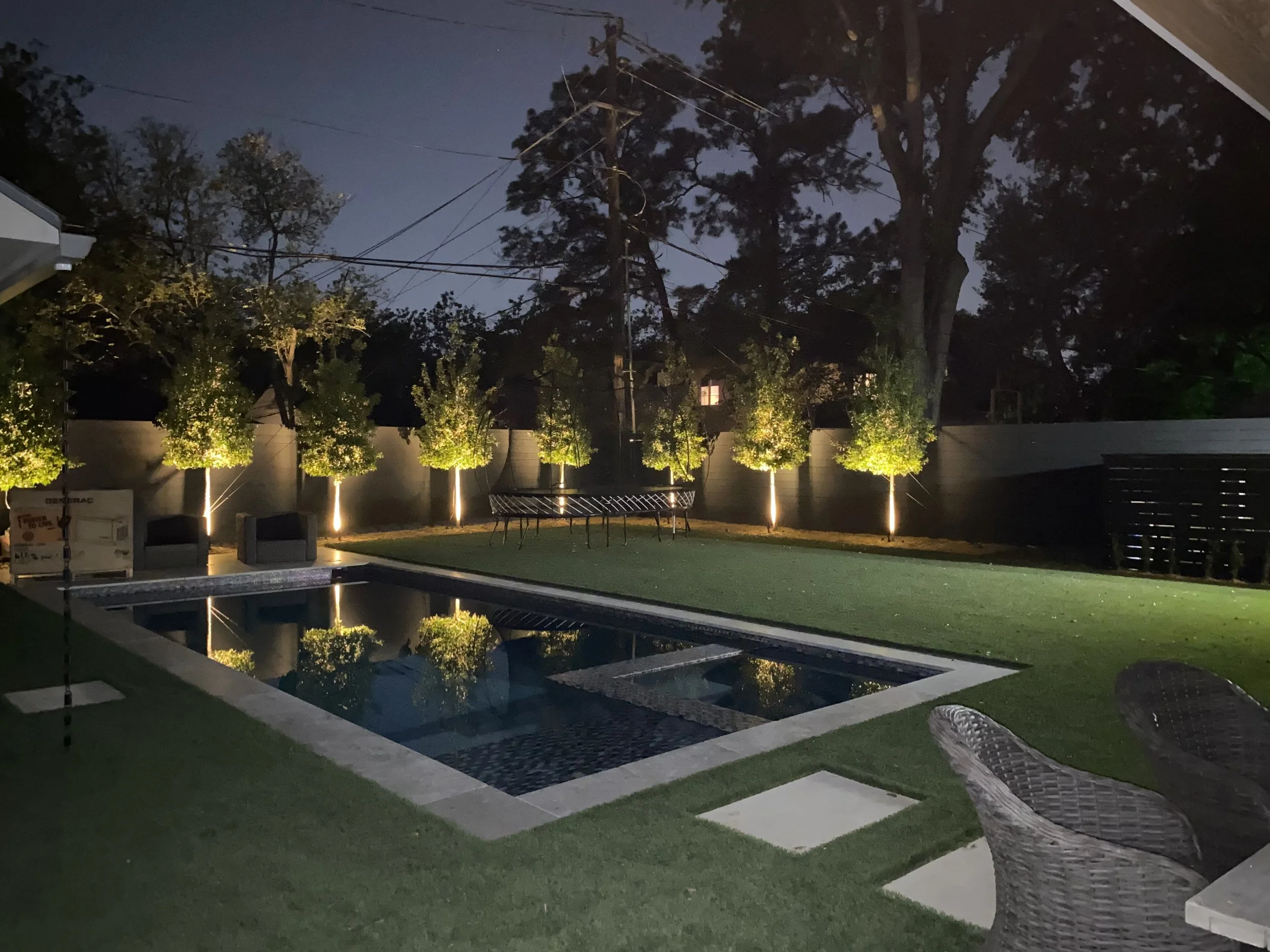 Nighttime backyard scene with an illuminated row of small trees along a fence, a swimming pool, outdoor furniture, and power lines in the background.