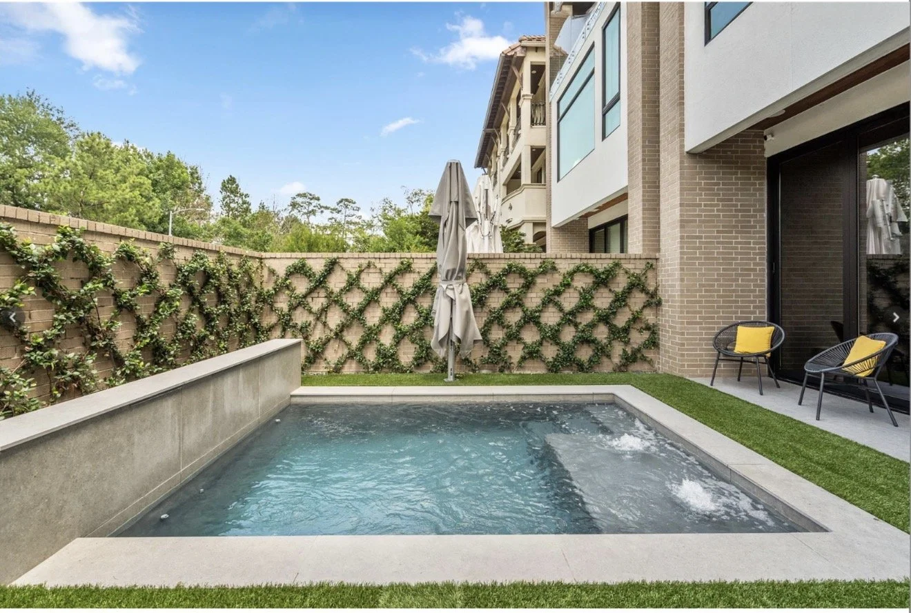 Small backyard pool with bubbling water, surrounded by a concrete border and artificial grass. Two black chairs with yellow cushions sit on a patio. A closed umbrella stands beside a vine-covered brick wall, with a multi-story apartment building in the background under a partly cloudy sky.