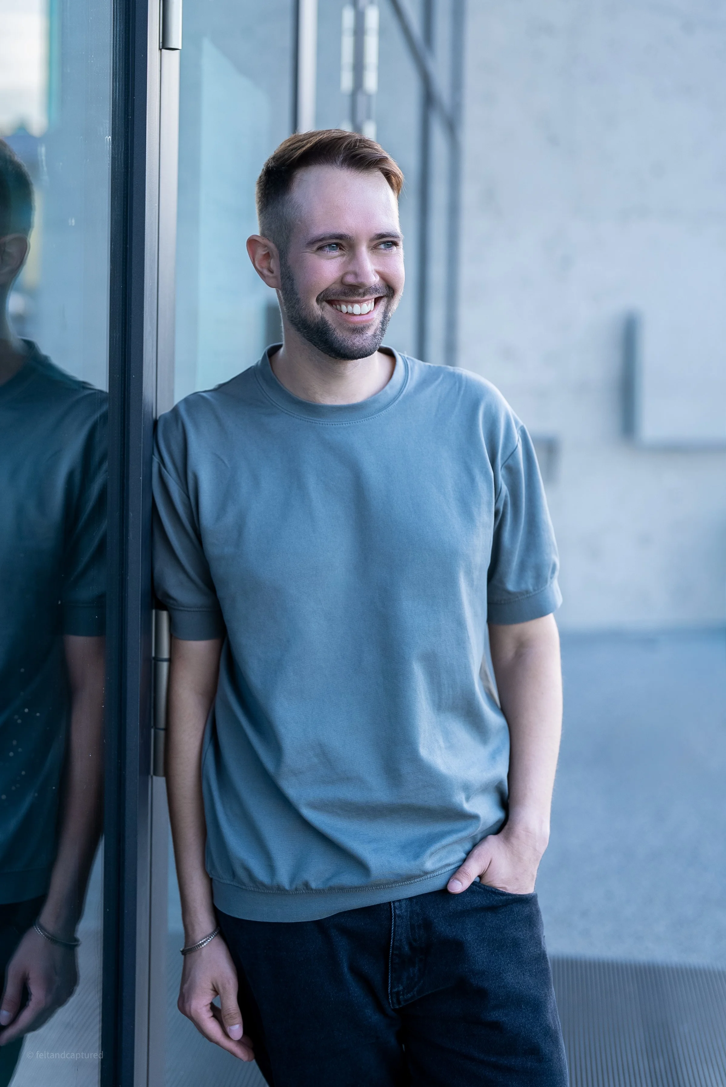 Ein junger Mann mit Bart und kurzen Haaren, trägt ein graues T-Shirt, lehnt an einer Glasscheibe und lächelt.