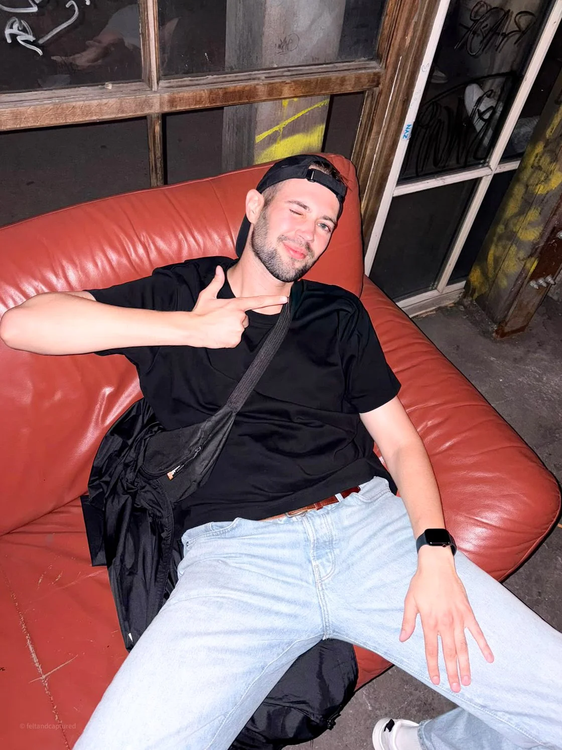 Junger Mann liegt entspannt auf einem roten Couch im Freien, trägt schwarze Kleidung, eine schwarze Cap, und macht eine Hand-Geste. Er scheint Spaß zu haben.