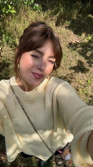 Junge Frau macht einen Selfie im Freien bei Sonnenschein, trägt einen cremefarbenen Pullover und eine Umhängetasche, schaut in die Kamera, während sie die Sonne genießt.