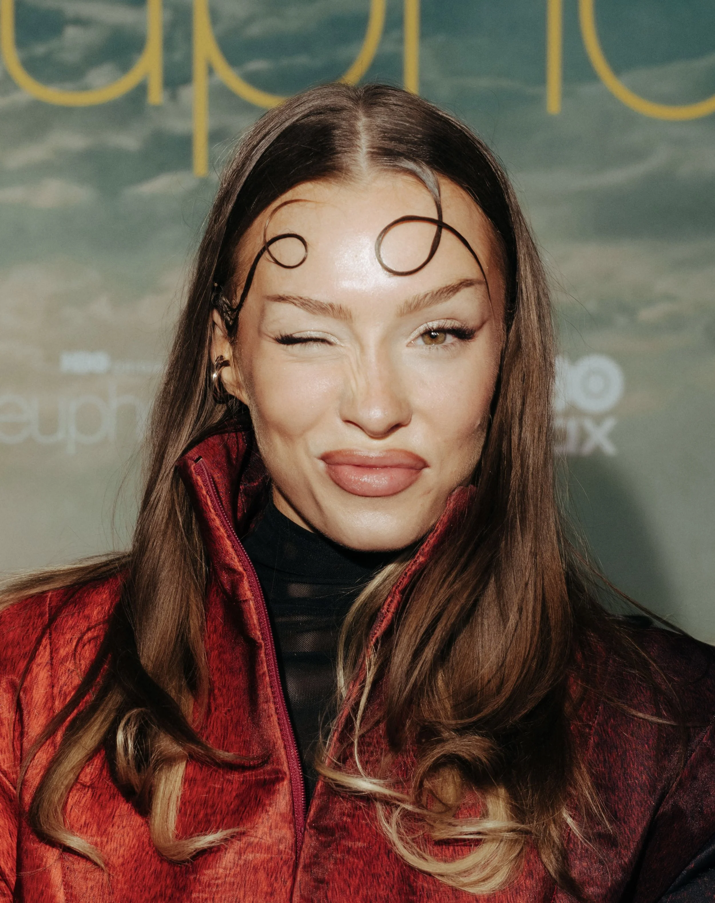 Eine junge Frau lächelt und macht ein lustiges Gesicht, trägt eine rote Jacke, hat lange vordere geflochtene Haare, braune Augen und trägt Ohrringe.