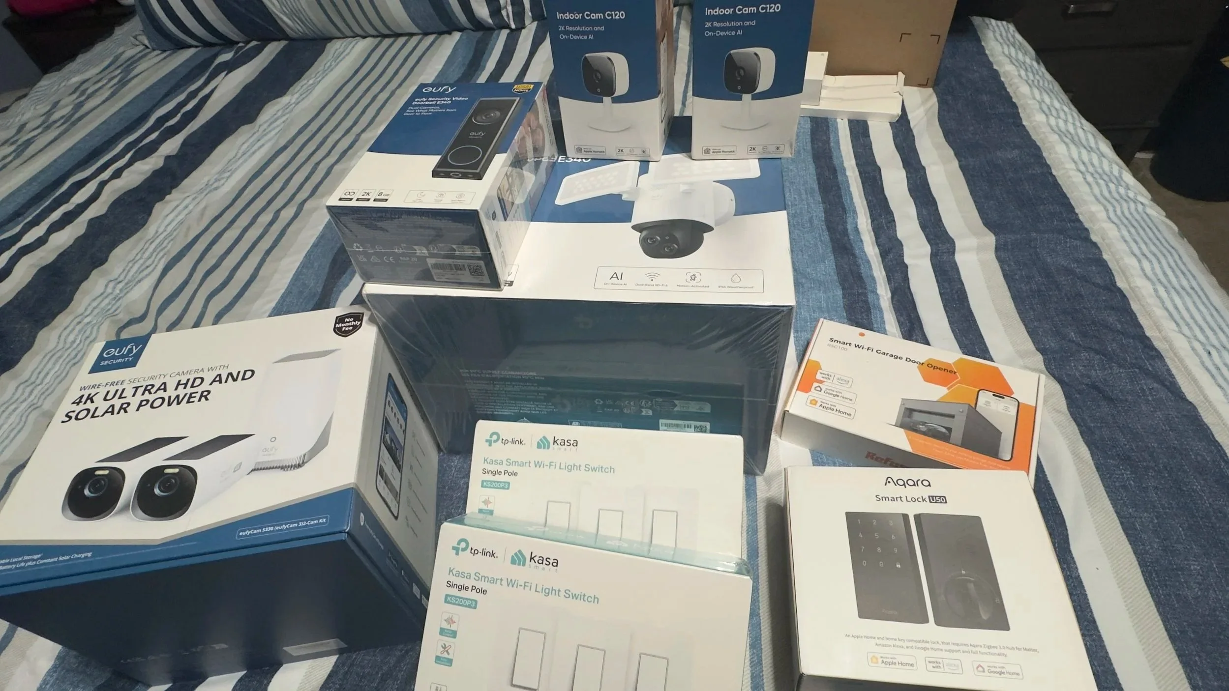 Various smart home security devices and accessories, including security cameras, a Wi-Fi light switch, a smart lock, and a garage door opener, arranged on a bed with a striped blue and white bedspread.
