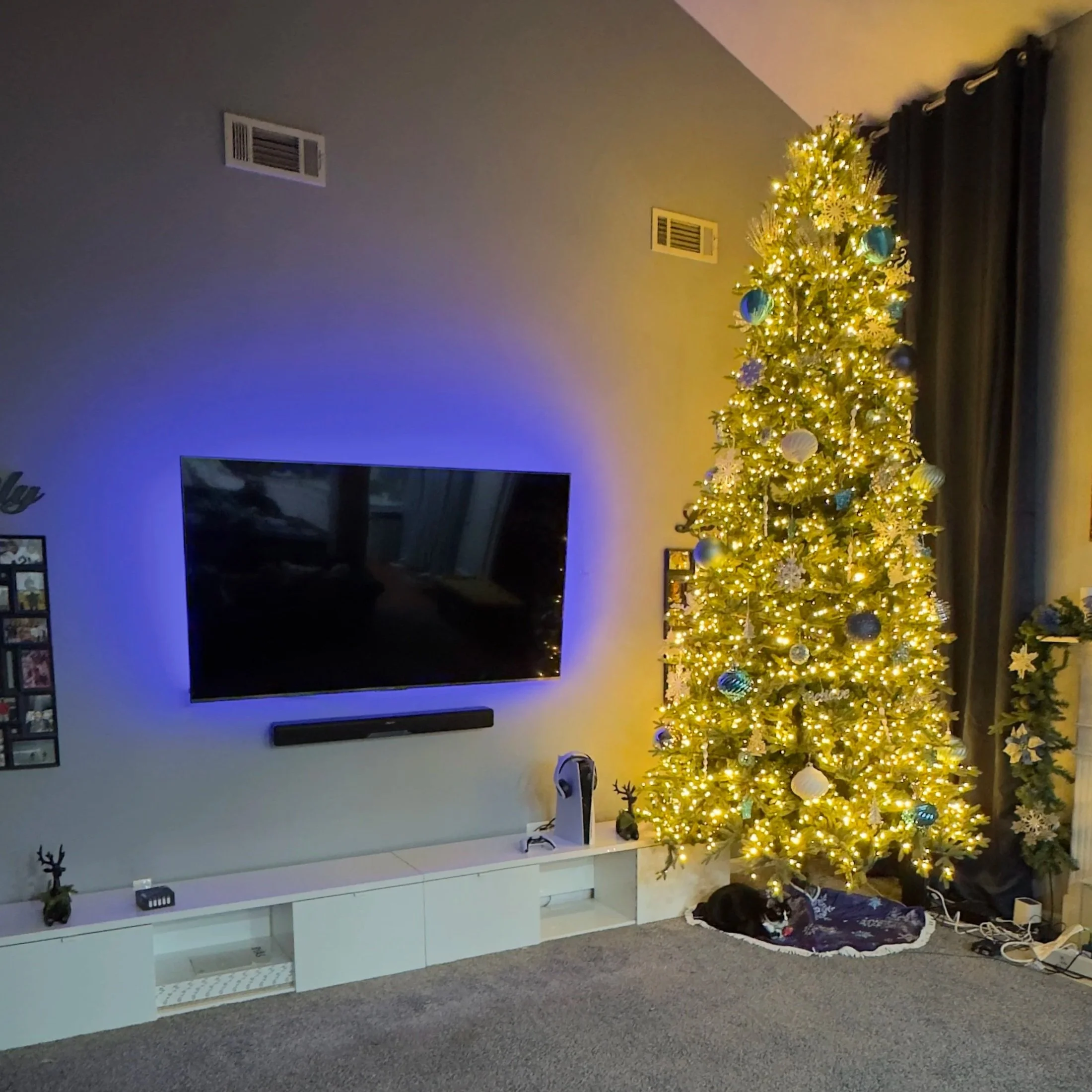 A decorated Christmas tree with white and blue ornaments and yellow lights in a living room next to a white entertainment center with a black TV. There are some electrical cords and a bag on the floor nearby.