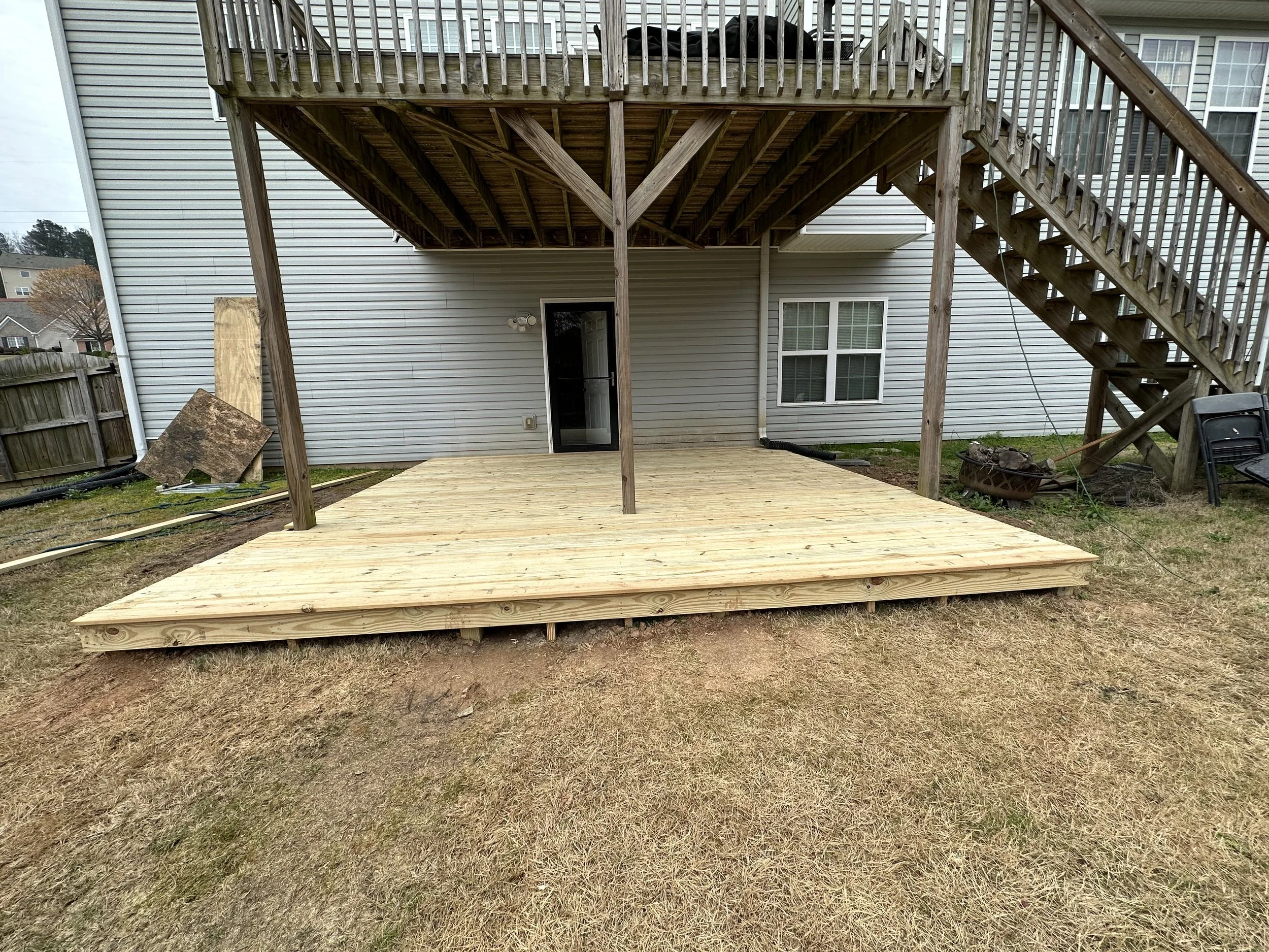 Wooden deck with stairs leading from upper to lower level, attached to a house with vinyl siding, a door and window, and construction materials on the ground.
