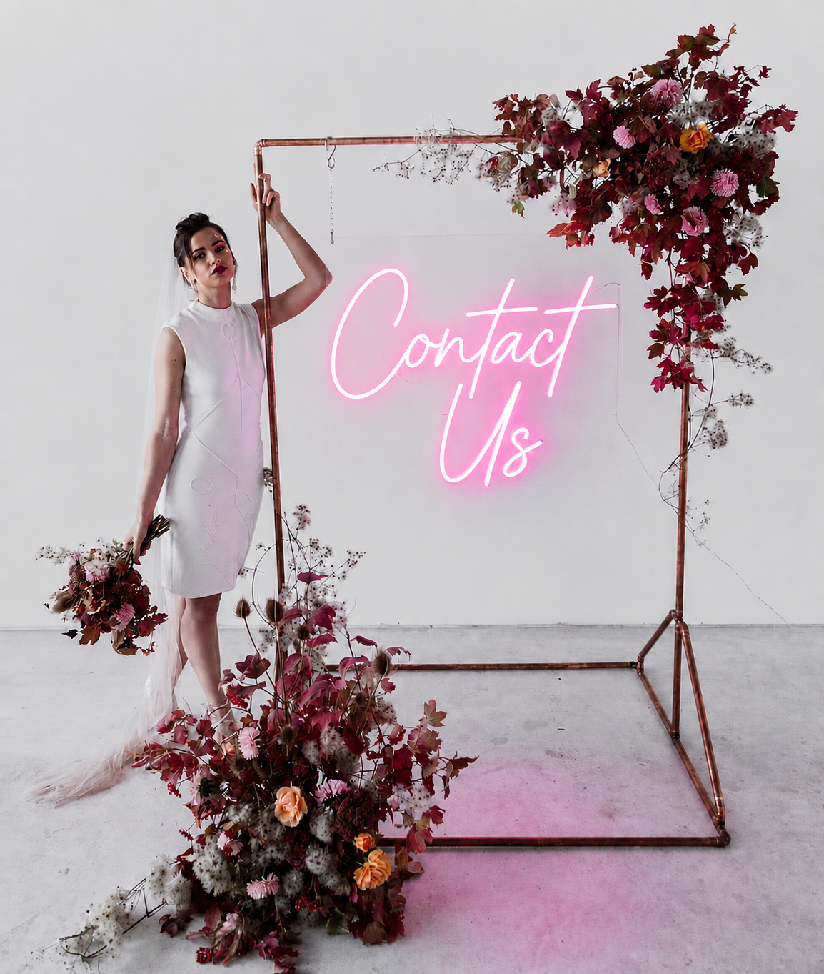 A woman in a white dress holding flowers stands next to a copper frame decorated with flowers and a pink neon sign that reads "Contact Us" in a minimalist setting.