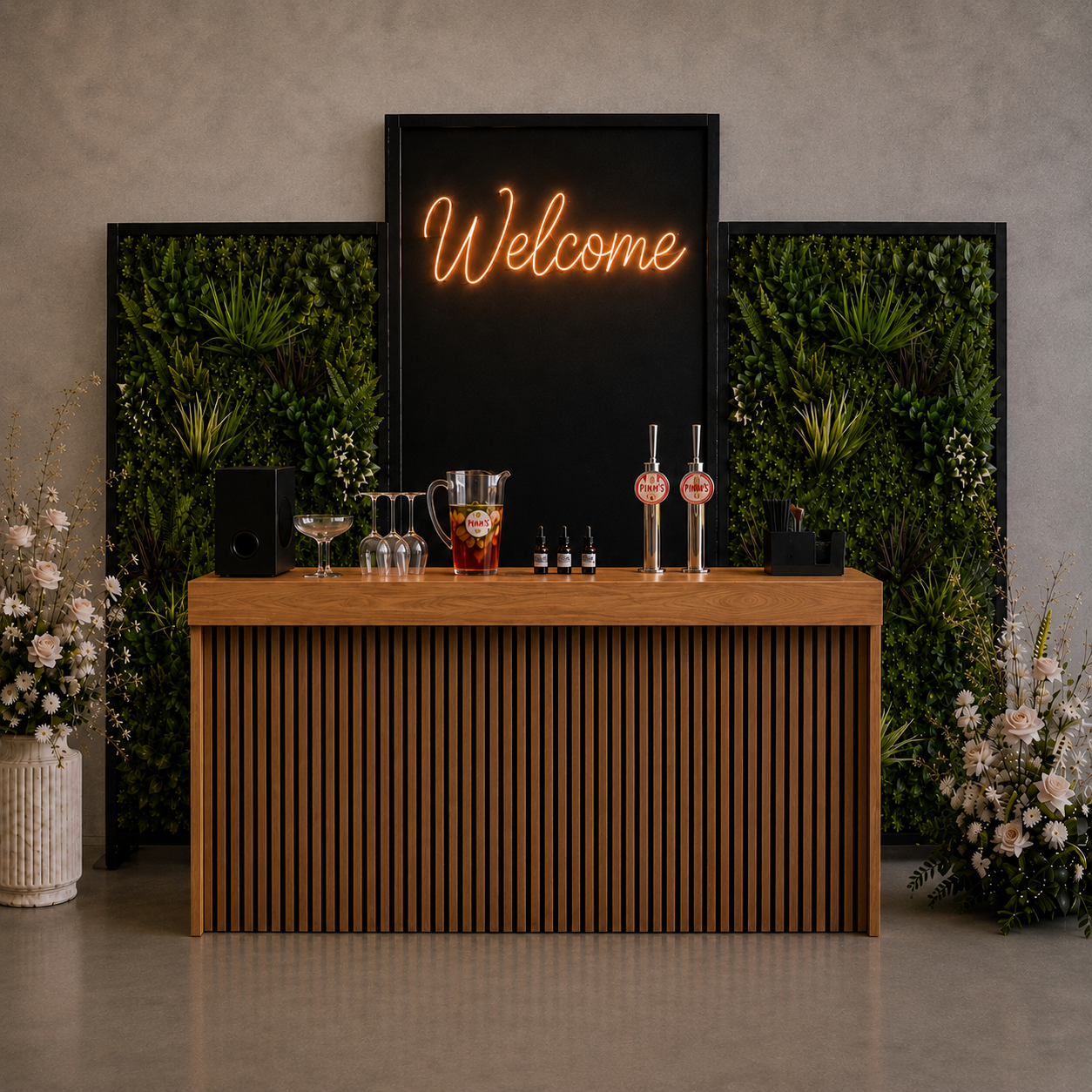 A reception bar with a wooden front, decorated with green plants on sides, a neon 'Welcome' sign on a black backdrop, and cocktail drink station with glasses, bottles, and dispensers.