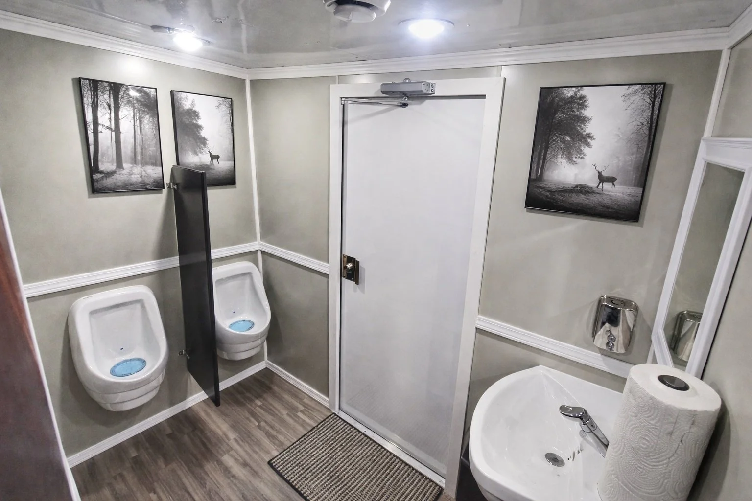 A clean men's restroom with two urinals separated by a black divider, a white sink with a paper towel roll on top, a mirror, and framed black-and-white nature photographs of trees and a deer on the walls.