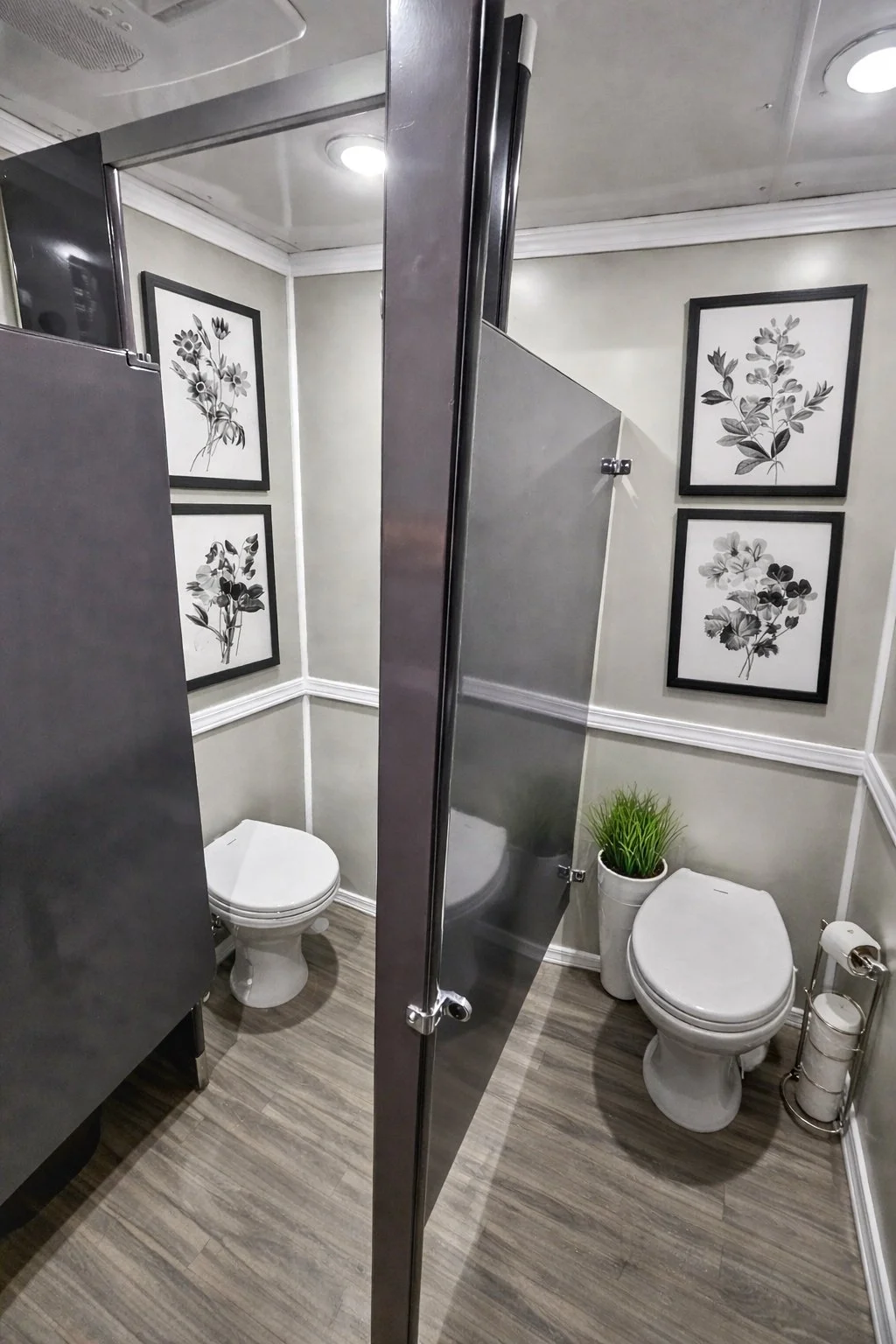 Public restroom with two toilets, black-frame botanical artwork on light gray walls, a potted plant, and a toilet paper holder.