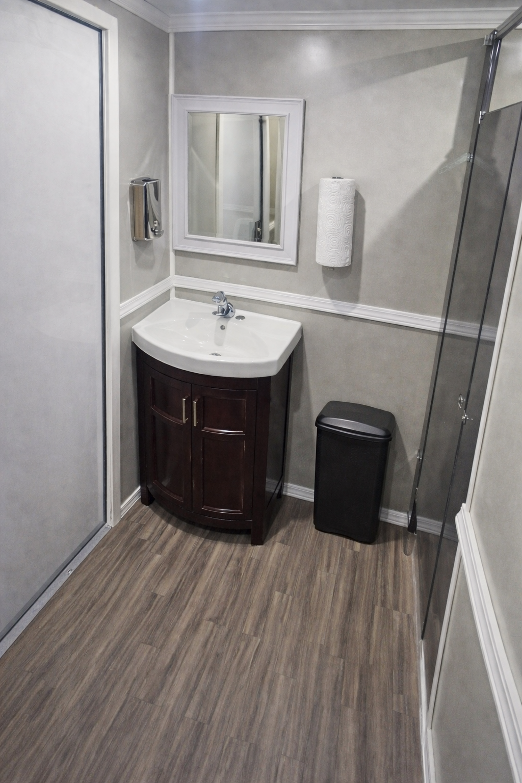 A small bathroom with a dark wood vanity sink, a mirror, a wall-mounted soap dispenser, a paper towel roll, a black trash can, and a small shower stall with a glass door.