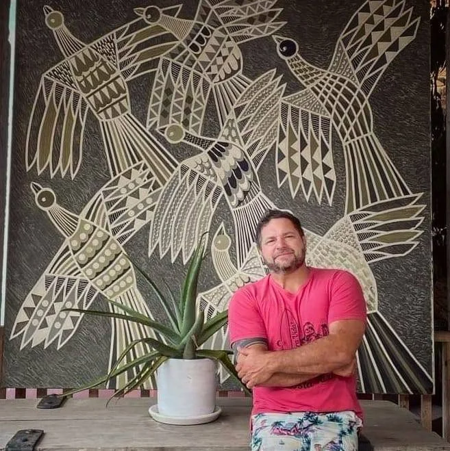 A man with a beard and short hair, wearing a pink t-shirt and patterned shorts, standing with folded arms in front of a wall art mural of stylized birds. In front of him, there's a potted plant with long, pointed leaves.